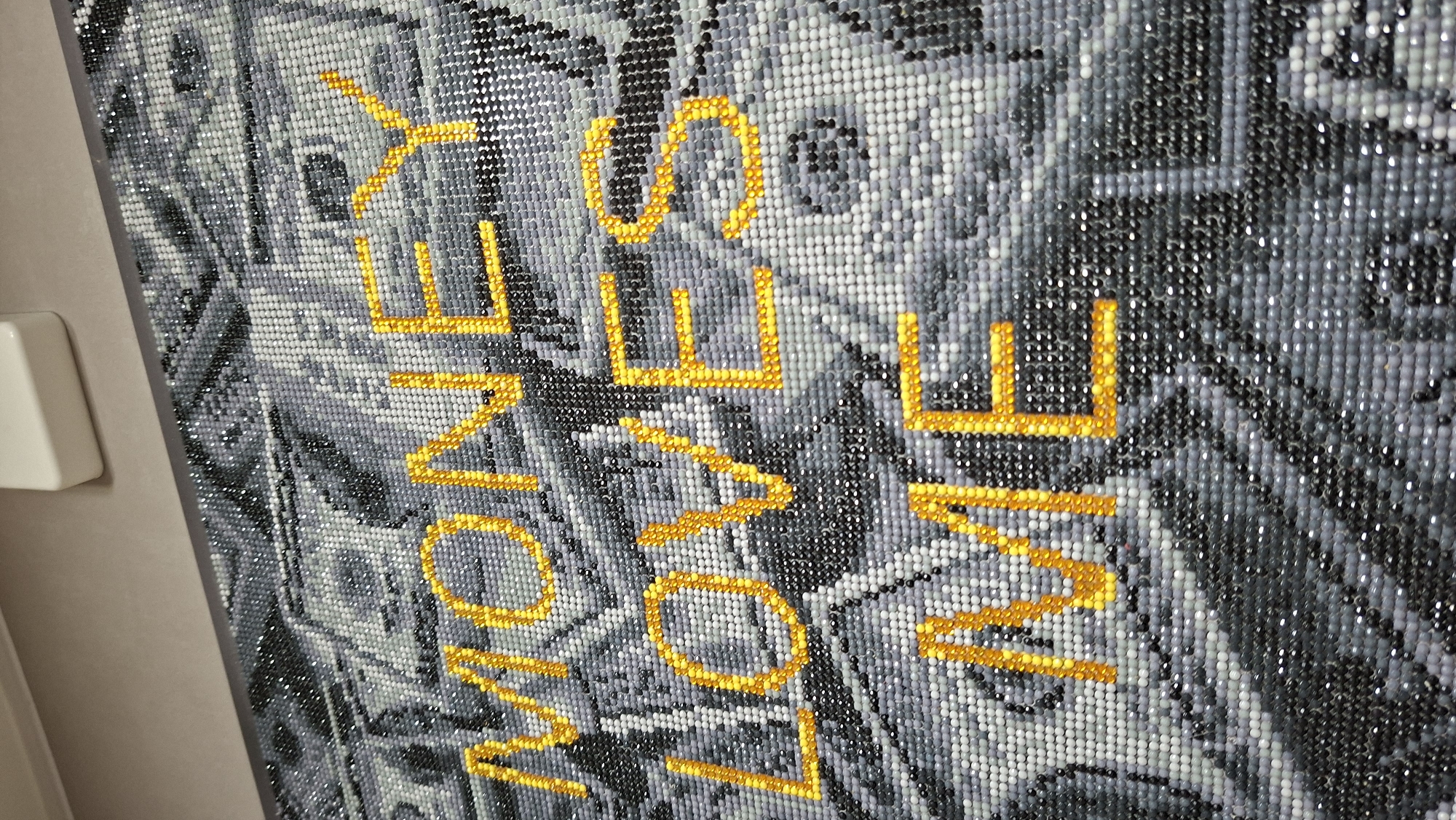 The image shows a diamond painting of what appears to be United States currency. The words "MONEY LOVE ME ALWAYS" are spelled out in gold lettering over the money imagery. The background is a mosaic of tiny, multicolored (mostly grayscale) beads or "diamonds." The overall effect gives the impression of a blurred image of money with the words in contrast. The painting is mounted and hung on a wall. A white electrical outlet or switch is visible to the left of the image. The location is unknown.