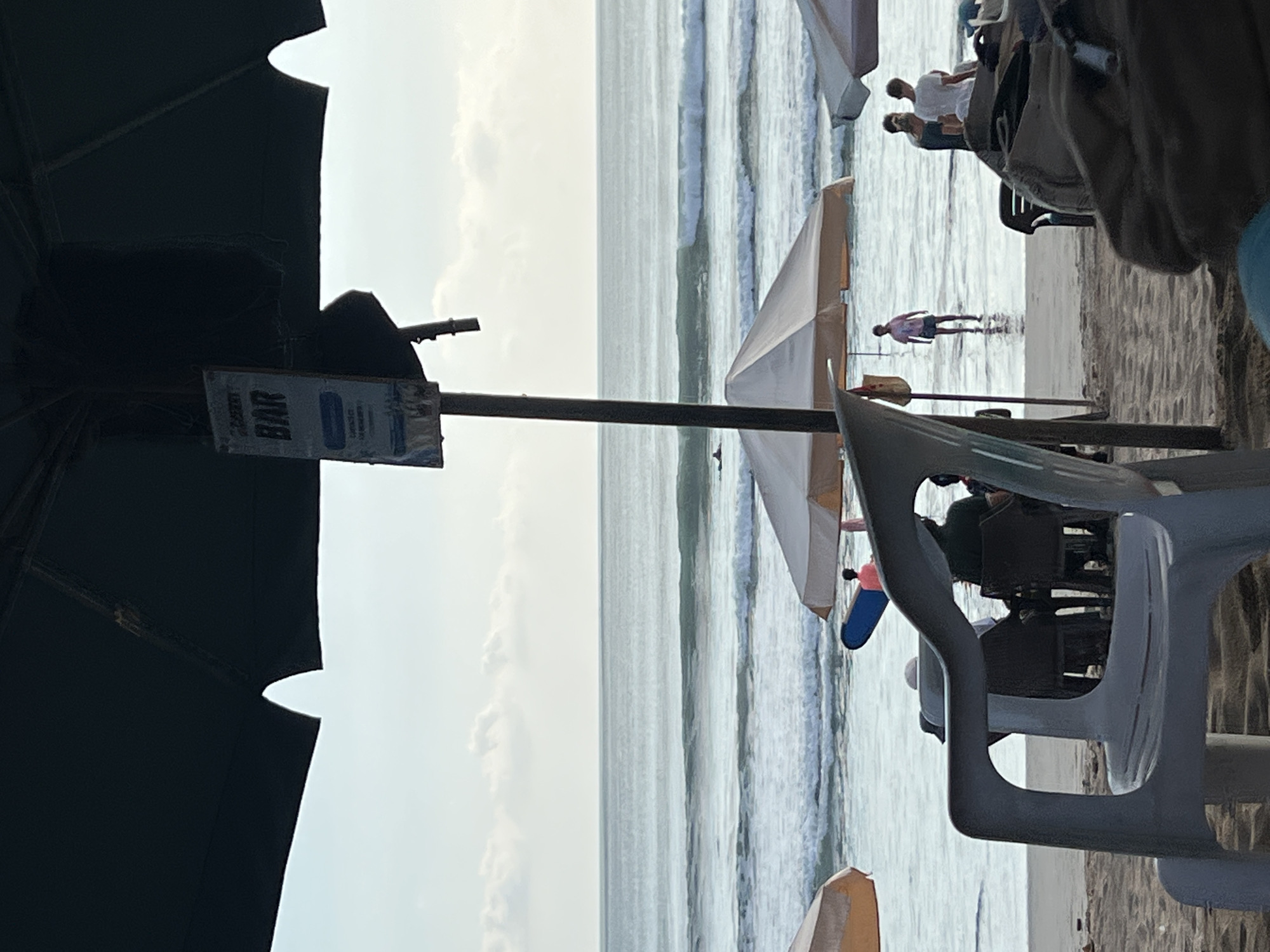The image depicts a beach scene in Kuta, Indonesia. In the foreground, a white plastic chair is positioned on the sand, partially obscuring the view of the beachgoers. Behind the chair, several individuals are seated at tables under beach umbrellas. Further back, the ocean is visible with gentle waves and a few surfers in the water. The sky is overcast with scattered clouds. A sign on a pole reads "BAR". The overall atmosphere suggests a relaxed day at the beach, with a bar and seating area set up for patrons.