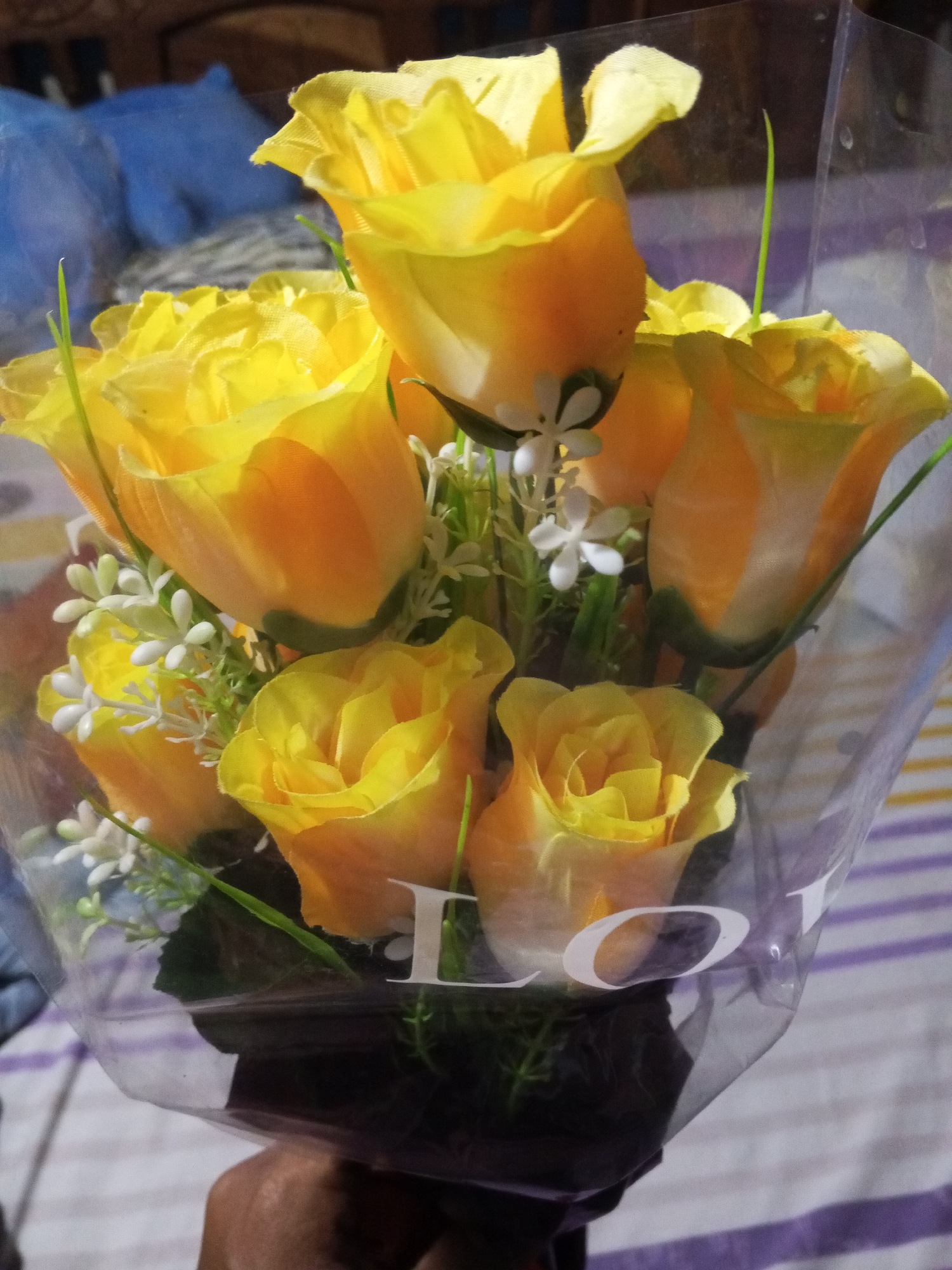 A bouquet of yellow roses is displayed in a clear plastic wrapper with the word "LO" visible near the bottom. The flowers are arranged with small white accent flowers and green stems. The background is a blurred pattern of light and dark lines. The city and specific location are not discernible from the image.