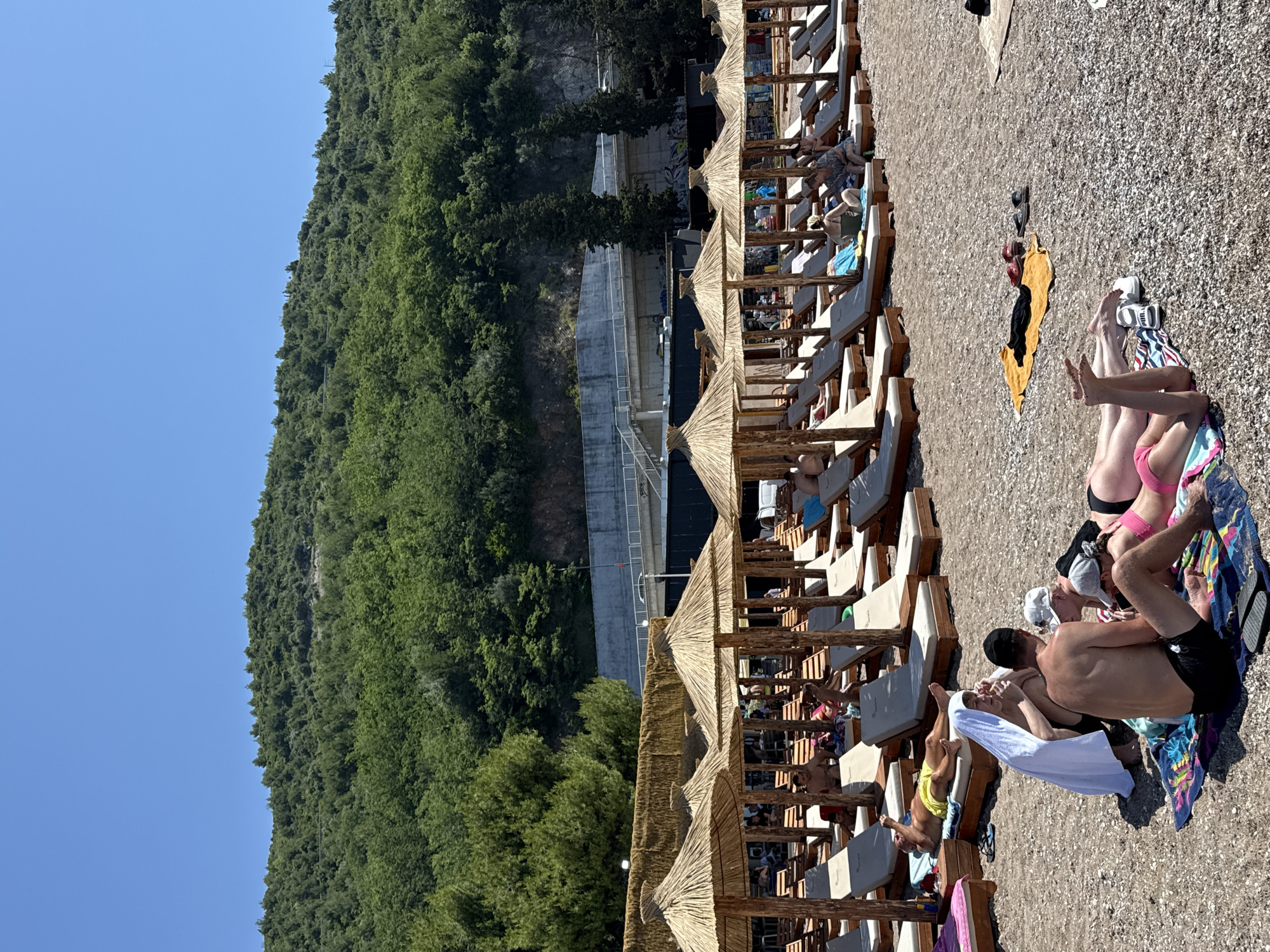 This image depicts a sunny day at a pebbly beach in Boreti, Montenegro, with a lush, green, tree-covered mountain rising in the background. The foreground shows numerous straw-thatched beach umbrellas arranged in rows, each shading multiple light-colored lounge chairs with wooden frames.
On the beach, several individuals and small groups are relaxing. In the immediate foreground, to the left, a person wearing pink swimwear is lying face down on a striped towel. To their right, a group of four people are gathered on a multicolored towel: two men (one shirtless in black shorts, the other in a white cap and T-shirt) and two women (one in a white towel wrapped around her head and shoulders, the other in a black cap and pink swimwear). The shirtless man faces the woman in the white towel, while the man in the white cap leans over. Further back and to the left, another man in yellow swimming trunks and sunglasses is lying on a lounge chair, with a white towel draped over a nearby chair.
Further into the midground, more people occupy the lounge chairs under the umbrellas, engaged in various activities like sunbathing or conversing. On the open pebbly beach, a yellow towel with black clothing, red sandals, and black flip-flops is laid out.
The midground features a dark building structure partially obscured by the umbrellas, with some visible graffiti. Above this, a concrete overpass or bridge with railings is visible, leading into the mountain. The mountain itself is densely covered in green trees and foliage, extending from the midground up to meet a clear, bright blue sky that dominates the upper portion of the image. The sunlight indicates a clear day.