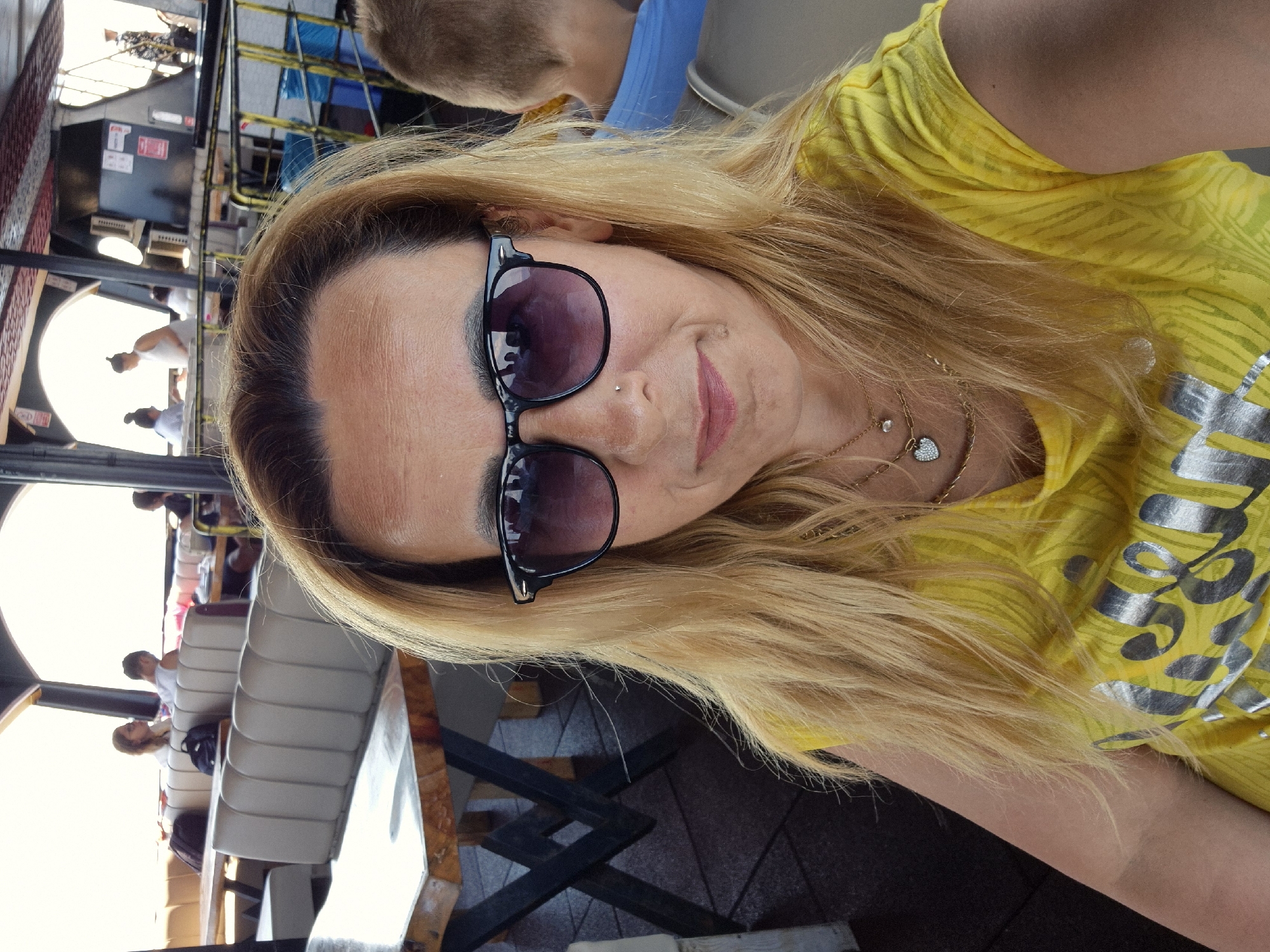 The image shows a woman with blonde hair, wearing sunglasses and a yellow t-shirt. She is in the foreground, and her face is turned towards the camera, with a slight smile. She is wearing a necklace with a heart-shaped pendant and a small diamond. In the background, other people are visible sitting on seats, possibly on a boat or a bus. There are also some tables and chairs, suggesting a seating area. The lighting suggests it could be daytime, with sunlight filtering in. The overall mood appears relaxed and casual. There is text on the woman's t-shirt, but it is partially obscured and difficult to read.