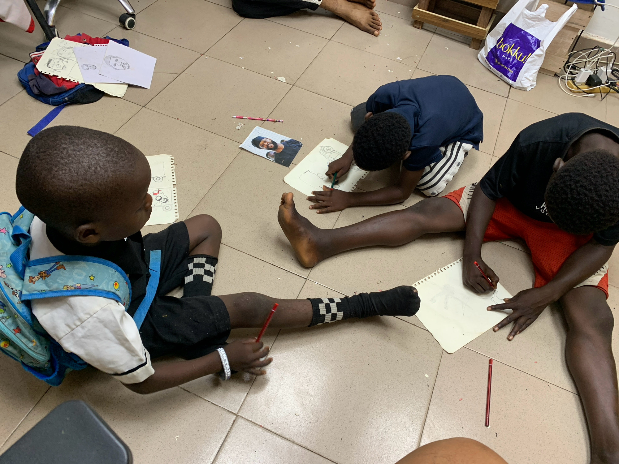 In Ado, Nigeria, three young Black boys are engaged in drawing on a tiled floor. One boy, wearing black shorts and socks with a checkered pattern, is sitting cross-legged on the right, drawing on a notebook with a red pencil. Another boy, in a blue t-shirt and striped shorts, is also drawing on a notebook in the center. A third boy, in the foreground with a blue backpack adorned with characters, is lying down and holding a red pencil. Various drawings and loose papers are scattered around them. A white shopping bag with the logo "bokku" is visible in the background, along with some power cords and a wooden structure. The scene suggests a casual, indoor setting, likely a home or community space.