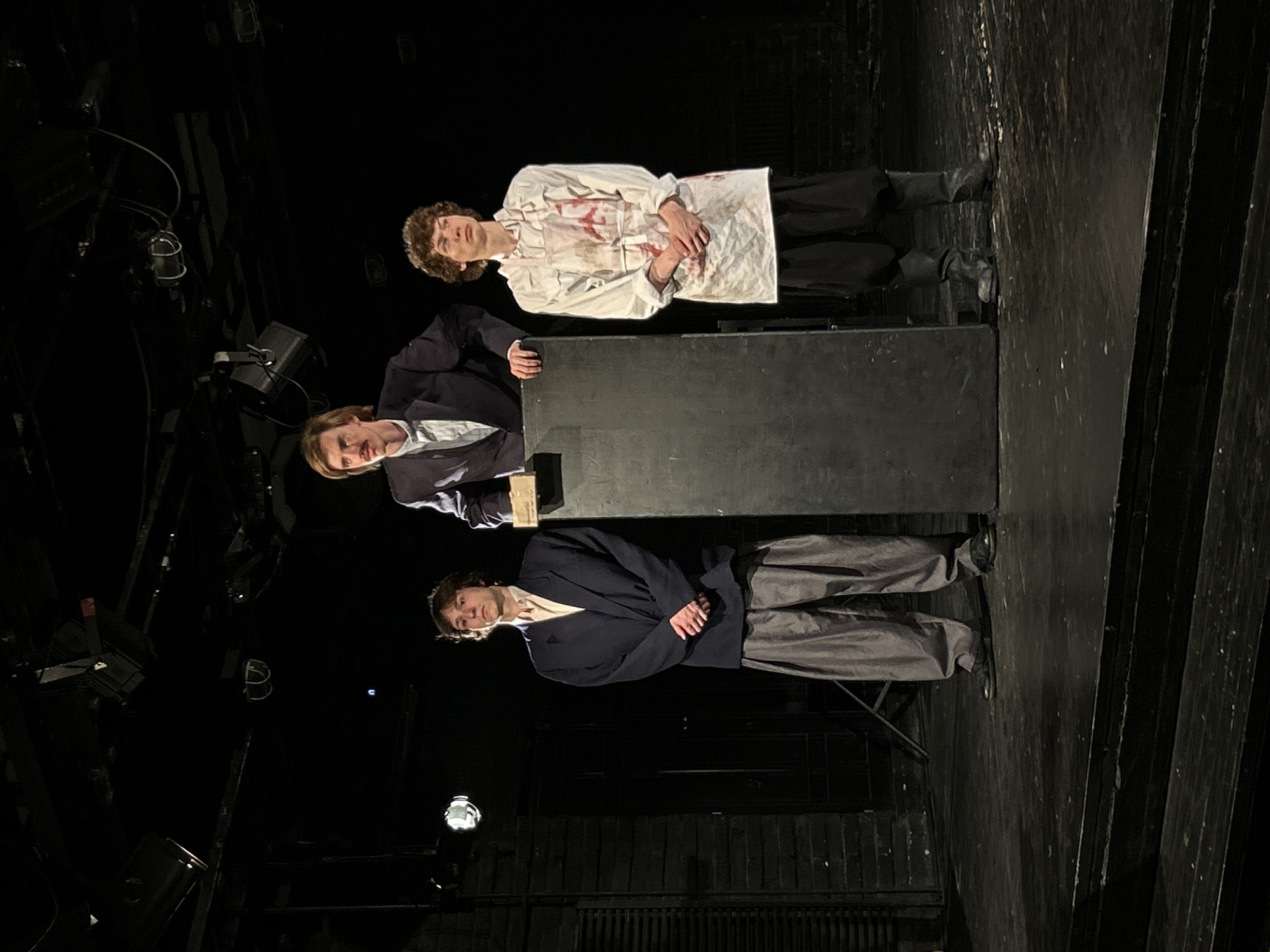 The image captures three males on a dimly lit stage within a performance venue in Unknown, Unknown. A man, appearing to be in his late 20s to early 30s with light brown hair and a mustache, stands centrally behind a tall, dark, rectangular podium. He is dressed in a dark suit jacket over a light-colored collared shirt. A small, light-toned object rests on the podium's upper surface.
To the viewer's left, a younger male, likely in his late teens or early 20s with dark, curly hair, stands facing forward. He wears an oversized dark blue blazer, a light-colored collared shirt, loose-fitting grey trousers, and dark shoes. His hands are clasped together at his waist.
To the viewer's right, another younger male, similar in age and with dark, curly hair, stands facing forward. He wears a long-sleeved white shirt and a white apron, both visibly stained with a significant amount of red liquid, consistent with simulated blood. He wears dark trousers and dark boots, with his hands also clasped at his waist.
All three individuals maintain an upright posture. The stage floor is dark, appearing to be constructed of wood, with two lower steps visible in the foreground. The background is predominantly dark, with stage lighting fixtures suspended from rigging visible in the upper frame and faint architectural elements, possibly dark brickwork or panels, discernible on the left side of the stage.