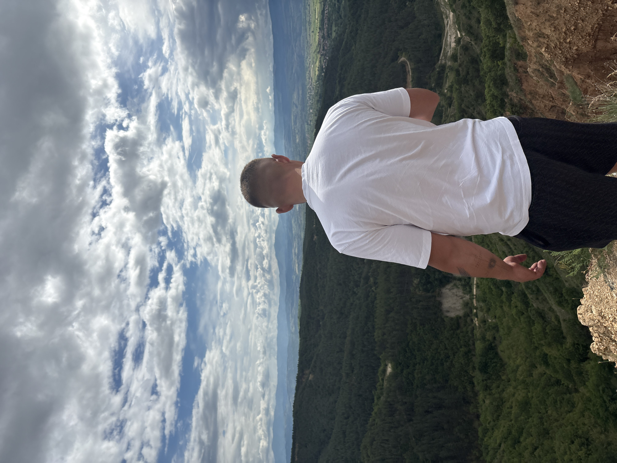 A man wearing a white t-shirt and dark shorts stands at the edge of a cliff, looking out over a valley and mountains. The sky is partly cloudy. The location is Porominovo, Bulgaria.