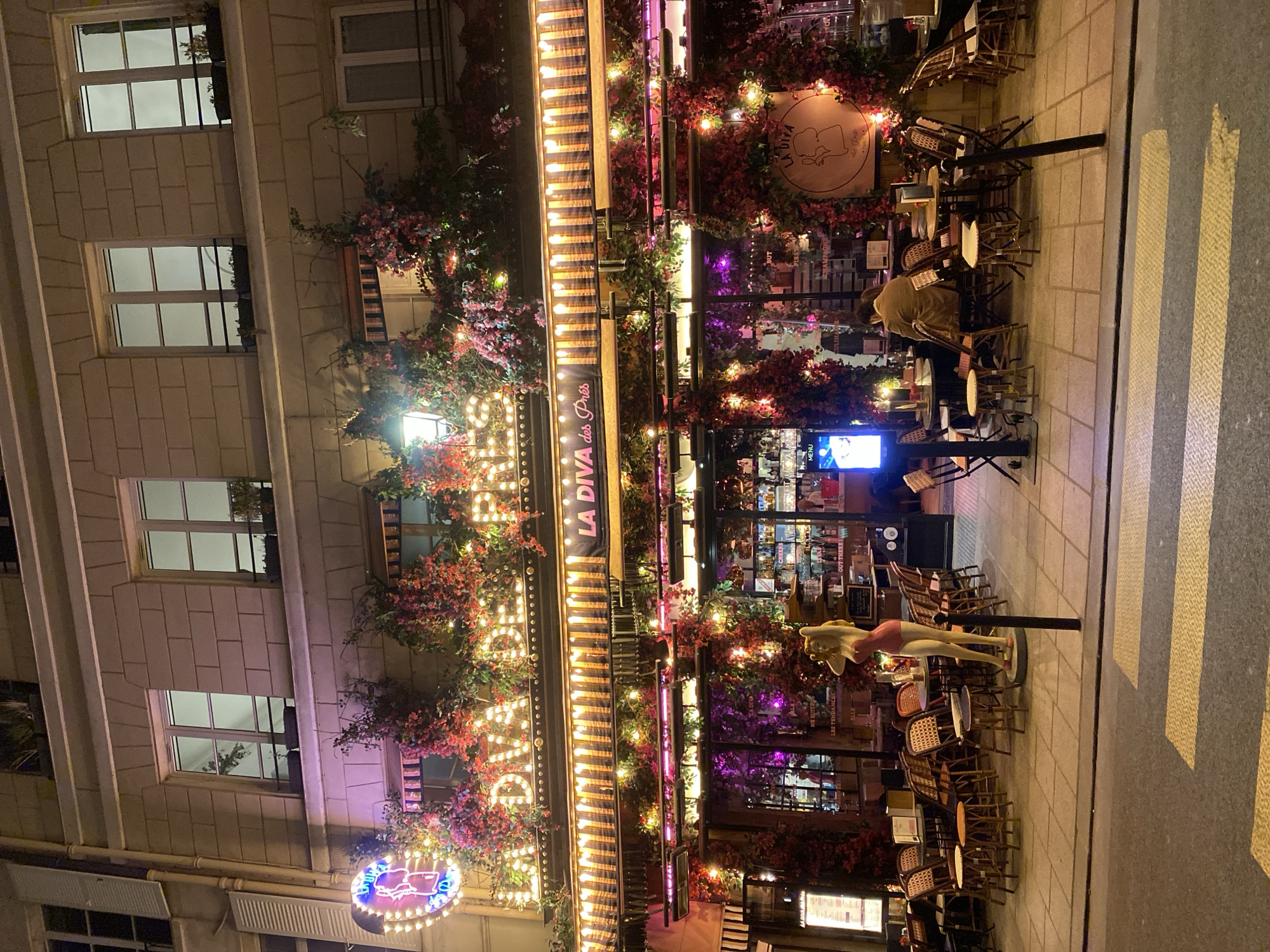 The image displays the brightly lit facade of an outdoor restaurant or cafe named "LA DIVA des Prés" at night, indicative of a bustling Parisian street scene. The building features light-colored, rectangular tiled upper floors with multiple windows, reflecting the ambient light.
The establishment's front is lavishly decorated with abundant artificial red and pink flowering plants or foliage, interwoven with numerous string lights that cast a warm, inviting glow. A large awning extends over the outdoor seating area, prominently displaying "LA DIVA des Prés" in illuminated lettering, further adorned with string lights. A circular sign bearing "LA DIVA" and a stylized graphic is visible on the facade, and a smaller blue and pink neon sign is seen towards the lower left.
The outdoor dining area features numerous bistro-style tables and chairs, many of which are stacked, suggesting either closing hours or a quiet evening. One person, wearing a light brown jacket, is seated at a table towards the right, facing away from the camera. A life-sized mannequin of a woman in a pink swimsuit stands upright near the tables. An electronic screen displaying "MENU" is visible in the background, near other signs with faint text like "CHAMPAGNE" and "LE TER...". The scene includes a paved sidewalk and a portion of a street with yellow road markings. The overall atmosphere is festive and lively due to the extensive lighting and decor, despite the relatively sparse activity.