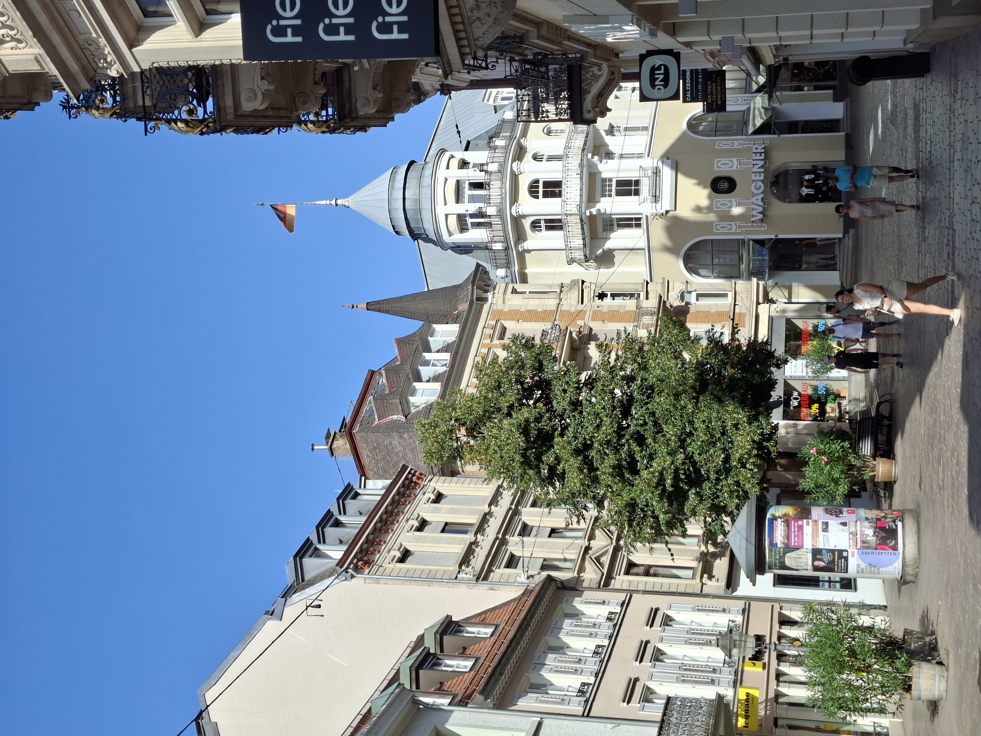 The image depicts a street scene in Baden-Baden, Germany, on a bright, sunny day. The architecture is characteristic of a European city, with multi-story buildings lining a cobblestone street. On the right side of the street, a building with ornate balconies and a German flag flying from its rooftop is visible. Adjacent to this building, a vertical sign displays the word "fielmann" in large white letters.
In the center of the image, a large green tree is prominent, its branches extending into the frame. Behind the tree, a building with a light yellow facade features a rounded turret with a conical roof, adorned with balconies. Below the turret, a shop with the name "WAGENER" is visible, its entrance featuring large glass windows.
Several people are walking along the street. In the foreground, a woman in a light-colored dress and a man in a blue shirt and shorts are walking away from the camera. To their left, a woman in a white dress and shorts is walking towards the camera. Further down the street, other individuals are seen walking.
The street is paved with cobblestones, and several potted plants and a bench are placed along the walkway, suggesting a pedestrian-friendly area. A cylindrical advertising pillar, covered in posters, stands in the middle of the street. The sky is clear and blue, indicating fair weather. The overall impression is that of a bustling urban environment on a pleasant day.