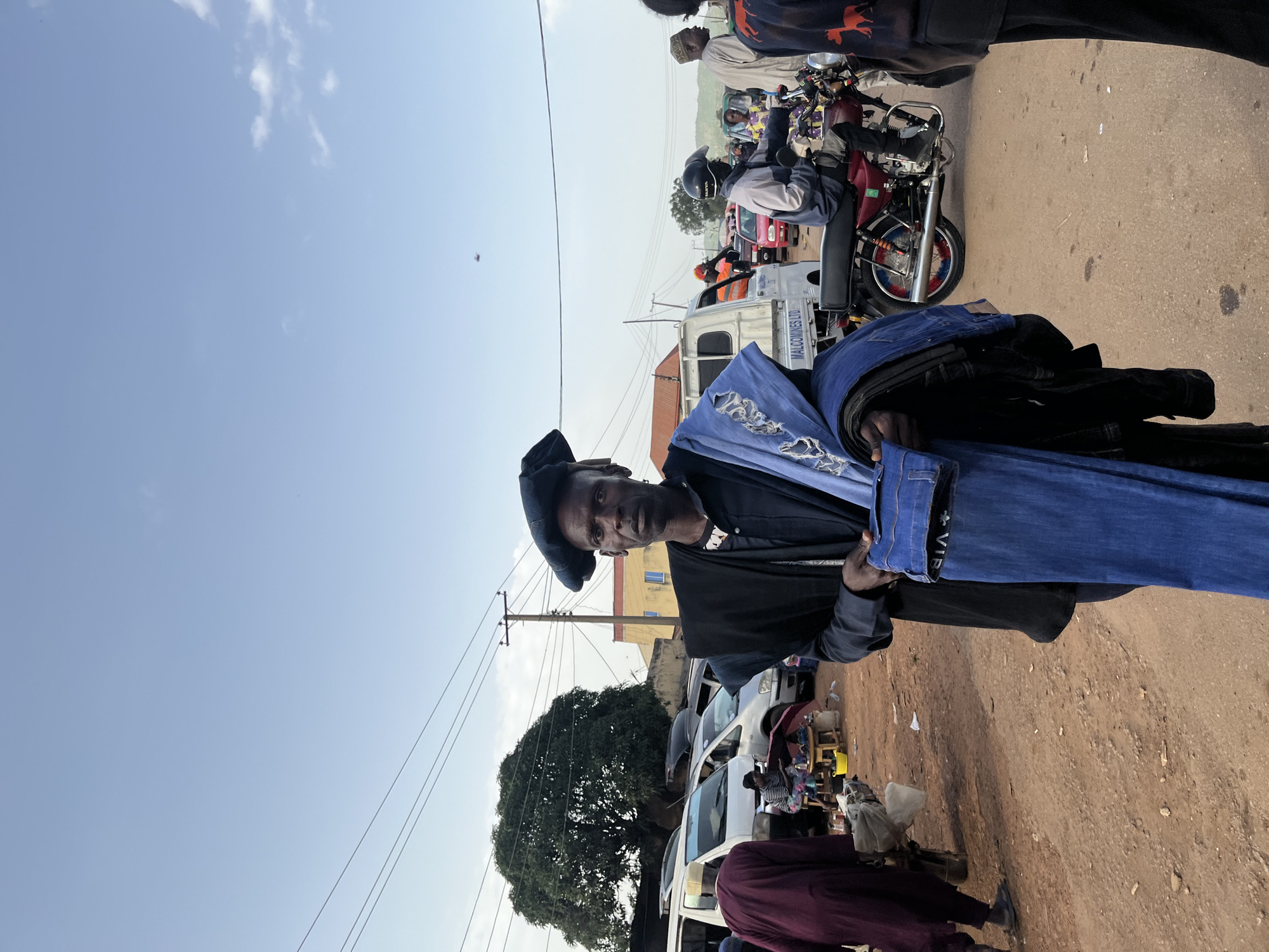 The image depicts a street market scene in Rafin Par, Nigeria. A man, likely a vendor, is prominently featured in the foreground, holding up several pairs of folded blue jeans. He is wearing a dark cap and a black garment. The scene is bustling with activity, with several motorcycles and a white truck visible in the background, suggesting transportation and commerce. Other people are present, some on motorcycles and others likely shoppers or vendors. The setting appears to be a dusty, outdoor market on a bright, clear day, indicated by the blue sky and sunlight casting shadows. The overall mood seems active and typical of a local market environment. A white truck in the background has text visible that reads "MALCOMINES LTD."