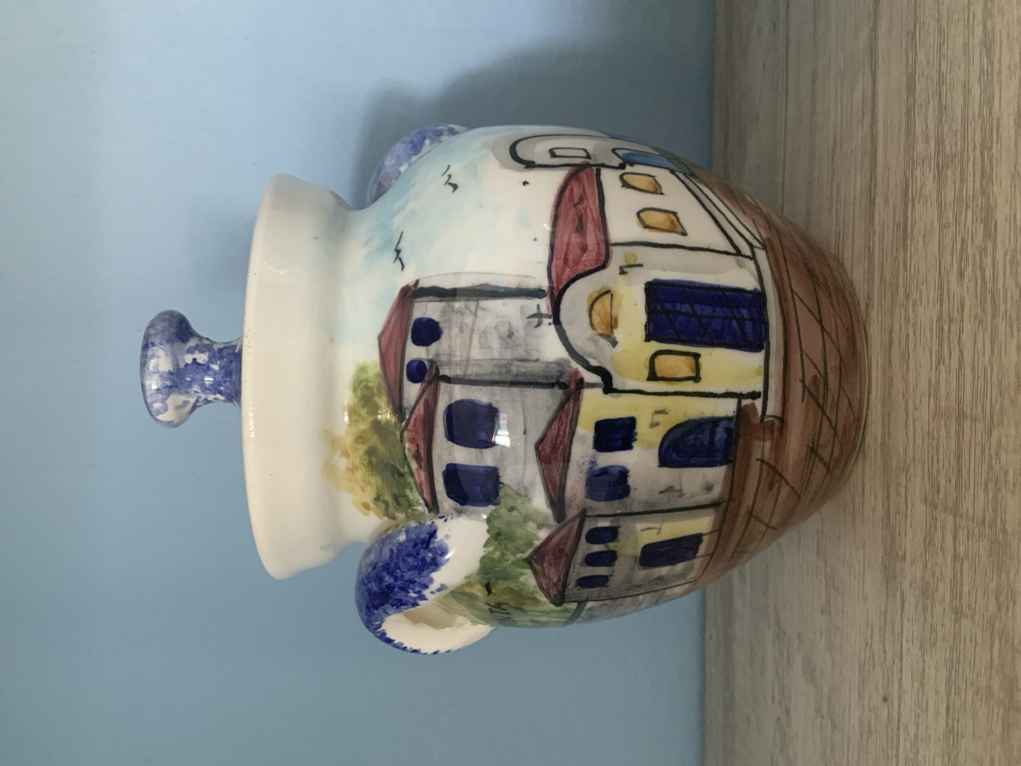 A white ceramic jar with a lid is displayed on a wooden surface against a blue background. The jar is decorated with a hand-painted scene of buildings with red roofs and blue windows, resembling a Mediterranean village. The scene also includes green trees and a light blue sky with two small black birds. The jar has two handles on either side, adorned with blue and white patterns. The lid has a similar blue and white patterned knob. The jar's base is painted with a brown pattern of horizontal and vertical lines. The overall style of the painting is naive and colorful.