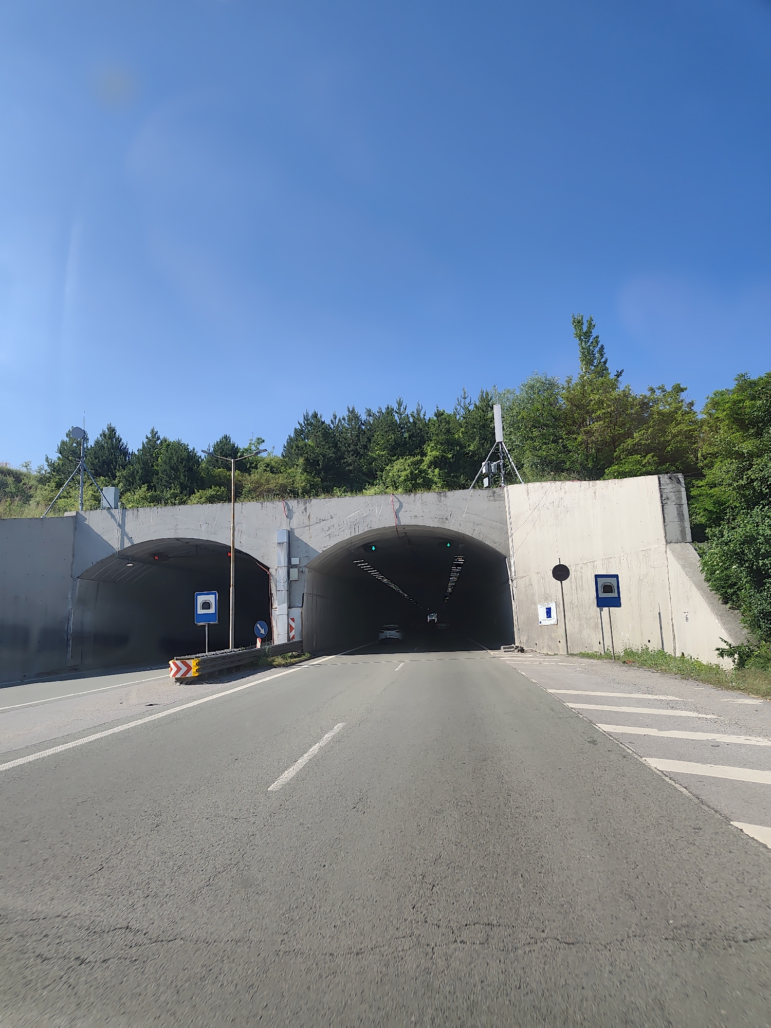 The image shows the entrance to a tunnel on a sunny day. The tunnel has two openings, side-by-side, with a concrete structure surrounding them. Road markings are visible on the asphalt, including white lines. There are traffic signs present, one indicating a tunnel entrance and another with a directional arrow. A yellow and red chevron warning sign is also visible on the left side of the road. Trees and vegetation are seen behind the tunnel entrance, and the sky is clear blue. The scene appears to be on a road leading into the tunnel.