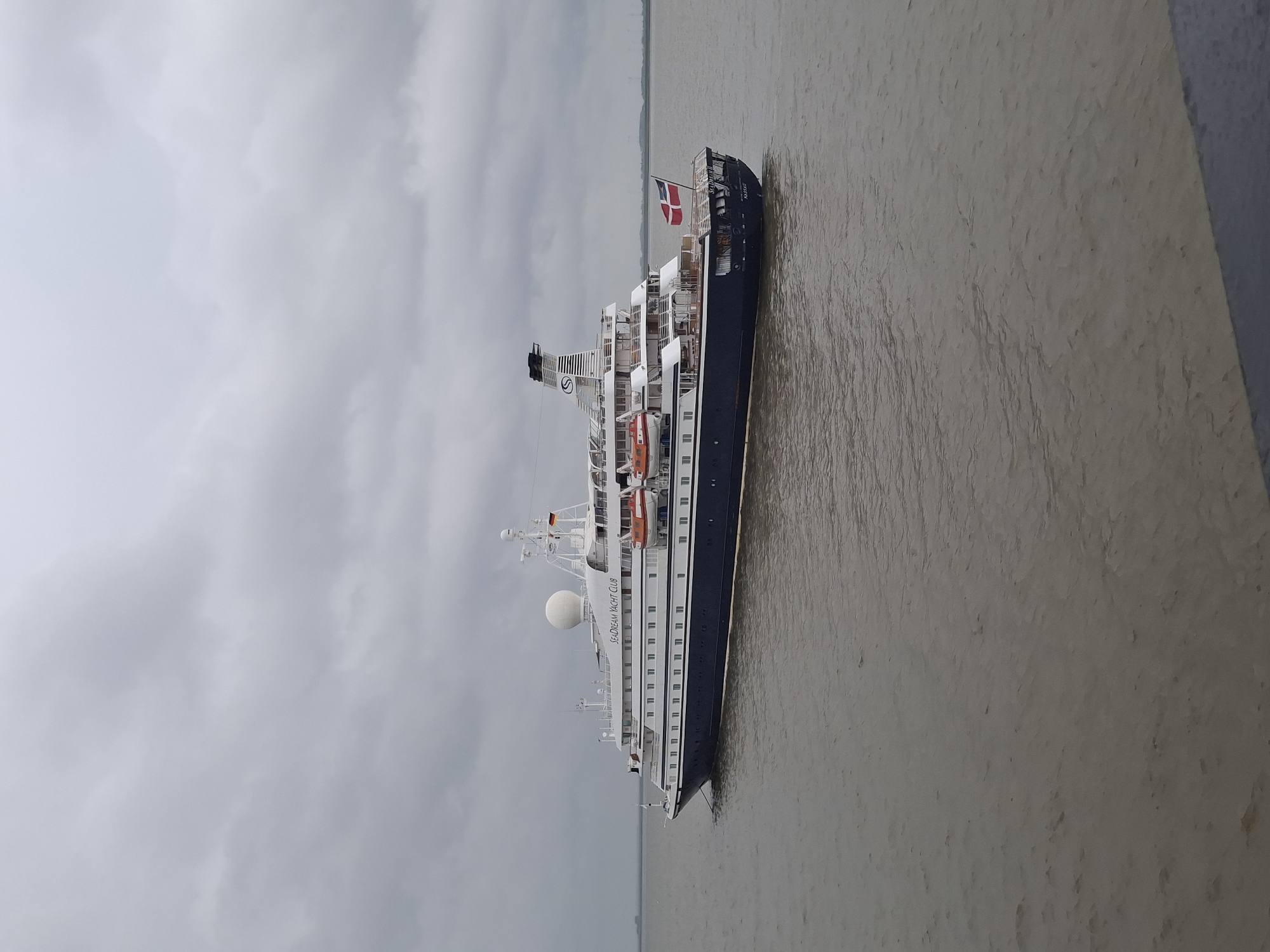 The image shows a ship, the SeaDream Yacht Club vessel, in the port of Brunsbüttel, Germany. The scene is characterized by overcast weather and a river, likely the Elbe, reflecting the dull light. The ship, with its black and white color scheme and orange lifeboats, is prominently displayed, with the Danish flag visible at the stern. The foreground includes a textured, light-colored surface.