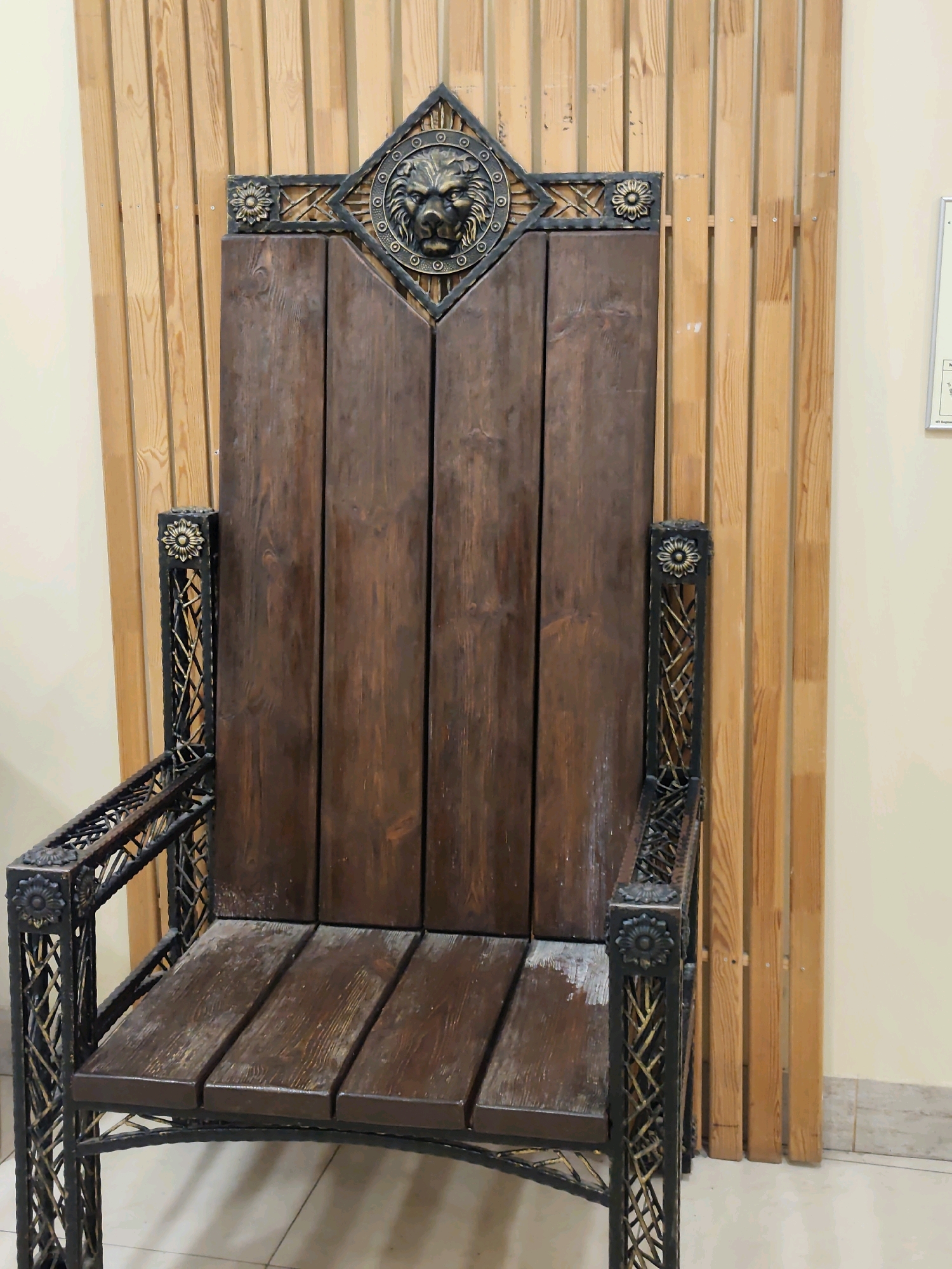 A regal wooden and metal throne-like chair sits against a backdrop of light-colored wooden vertical planks. The chair has a dark wooden seat and backrest, divided into segments by vertical lines. The armrests and frame are constructed from black metal with ornate, gold-colored floral accents. The top of the chair features a decorative metal crest with a lion's head in the center, surrounded by circular patterns and smaller floral embellishments. The chair appears to be an exhibit or decorative piece in a public space. The image does not provide any specific information about its location in Kazan, Russia, or any people or actions.
