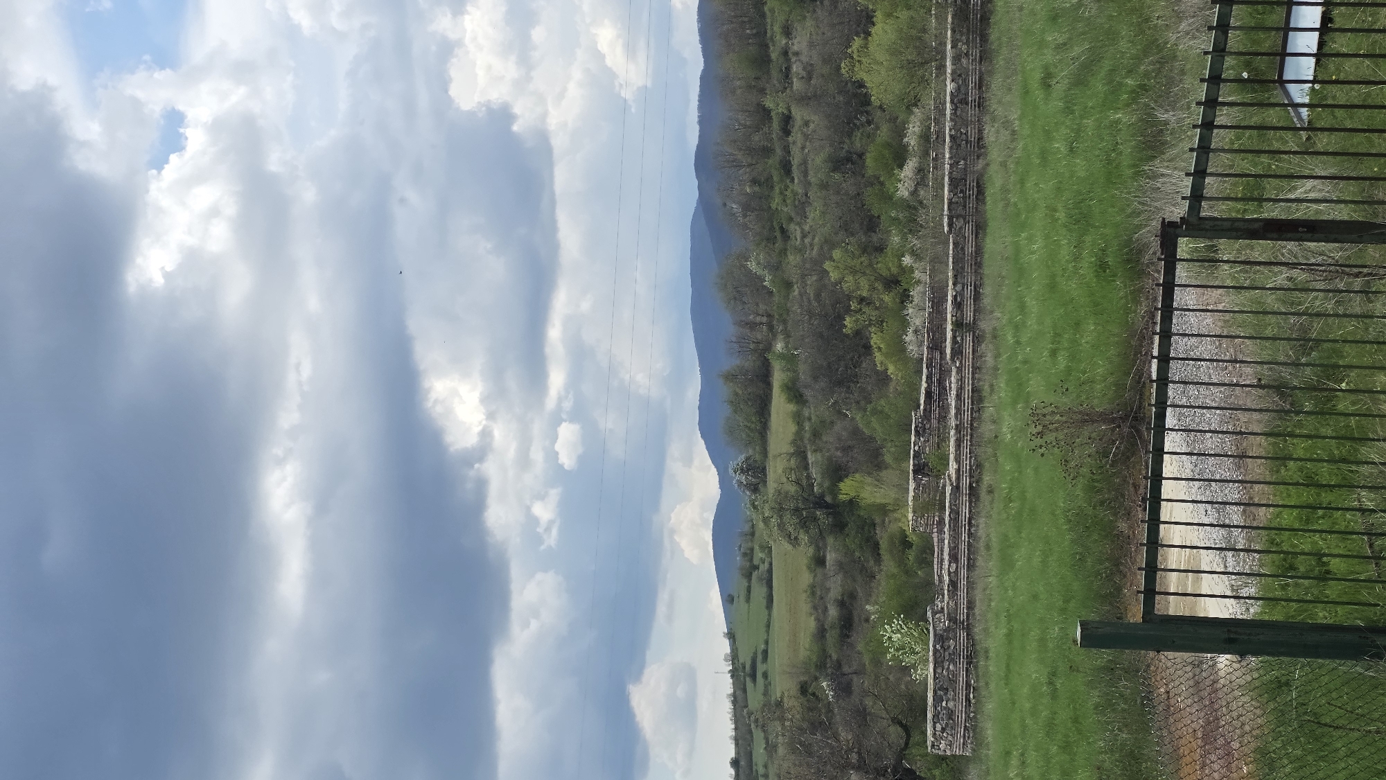 A vibrant landscape image, likely taken in Drenkovo, Bulgaria, showcases a rural mountainous scene under a partly cloudy sky. The upper portion of the frame is dominated by a bright sky featuring large, dynamic white and grey cumulus clouds, with patches of clear blue sky visible between them.
A range of forested mountains or hills stretches across the middle ground, displaying various shades of green foliage. Below the distant mountains, a valley unfolds with expansive, lush green fields. Scattered trees, some with white blossoms, are interspersed throughout the landscape, adding texture and lighter tones.
In the mid-ground on the right, ancient-looking stone or brick structures, possibly remnants of historical buildings or terraced foundations, are partially covered by vegetation. Closer to the viewer, along the right edge of the foreground, a dark green metal fence with vertical bars stands, partially obscuring a gravel path or dirt road. Faint utility lines are visible, vertically traversing the sky.
The scene is brightly illuminated, indicating daytime, likely morning or afternoon. The overall atmosphere is peaceful and serene, with no human or animal activity explicitly depicted. No visible text is present in the image.