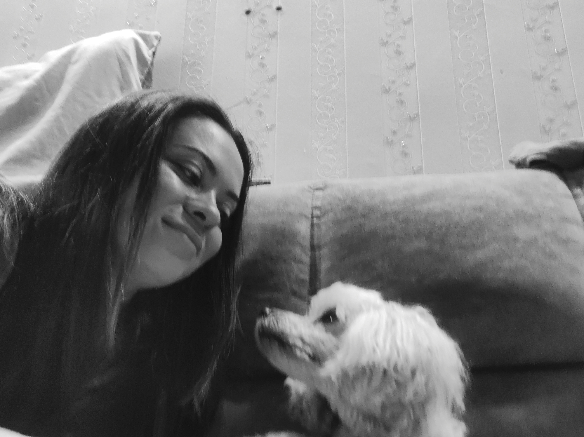 In this black and white, medium-angle, selfie-style photograph, a young woman with long, dark hair is looking down and smiling at a small, fluffy, white dog. The woman's face is in profile, with her eyes slightly closed and her cheeks puffed out. She appears to be lying down or reclining. The dog is positioned to the right of the woman, looking up at her. Its head is tilted slightly, and its fur is curly and soft. The background consists of a textured, striped wallpaper and a piece of furniture, likely a sofa or a couch, with a quilted pattern. A draped fabric, possibly a blanket or sheet, is visible on the left side of the image. The scene appears to be indoors, in a residential setting. There are no specific landmarks or indicators that would suggest a particular location within the city of Chaikovsky, Russia.