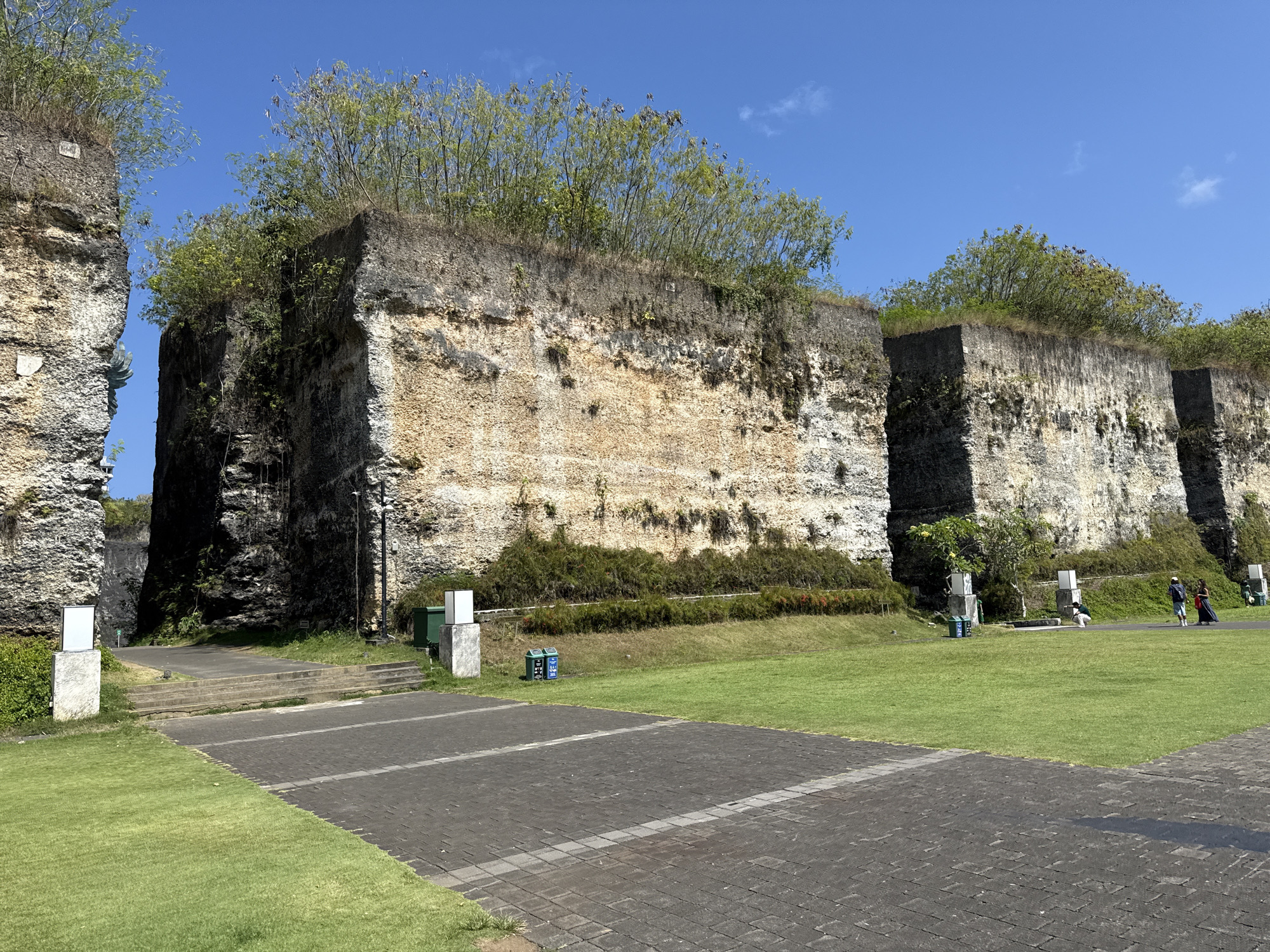 The image shows a public park in Kuta Selatan, Indonesia. The park features high, artificial stone cliffs with trees and vegetation growing on top. A paved brick pathway and manicured lawn are in the foreground. There are several people walking in the park in the background. There are also small, white information stands, trash cans, and a green utility box. The sky is clear and blue.