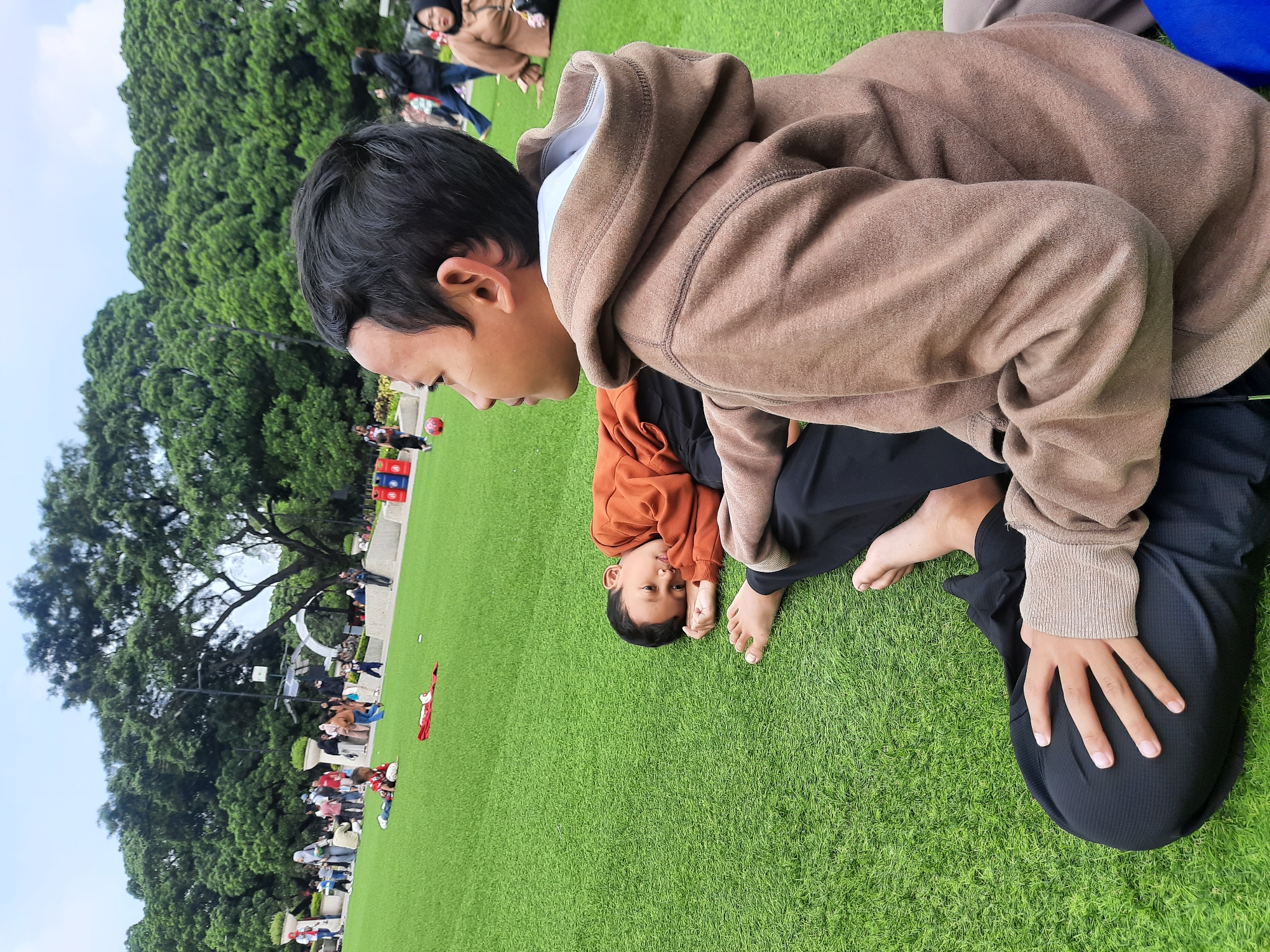 Two boys are sitting on a bright green, artificial turf lawn in a park. The boy in the foreground is on the right, facing to the left and slightly upwards. He has dark hair and is wearing a brown hooded sweatshirt over a white shirt and black pants. His right hand is resting on his left knee, which is bent. To his left, a younger boy is lying on his side, facing towards the foreground. He has dark hair and is wearing an orange long-sleeved shirt and black pants. His head is propped on his hands, and he appears to be resting or looking towards the other boy.
In the background, there are more people walking and playing on the lawn, and further back, there is a dense line of large, mature green trees under a cloudy sky. There are also some playground-like structures and red and blue bins visible in the distance. The overall impression is of a public park, likely in a city, with recreational activities taking place. Based on the context of Bandung City, Indonesia, this scene could be at a popular park like Taman Film or a similar recreational green space.