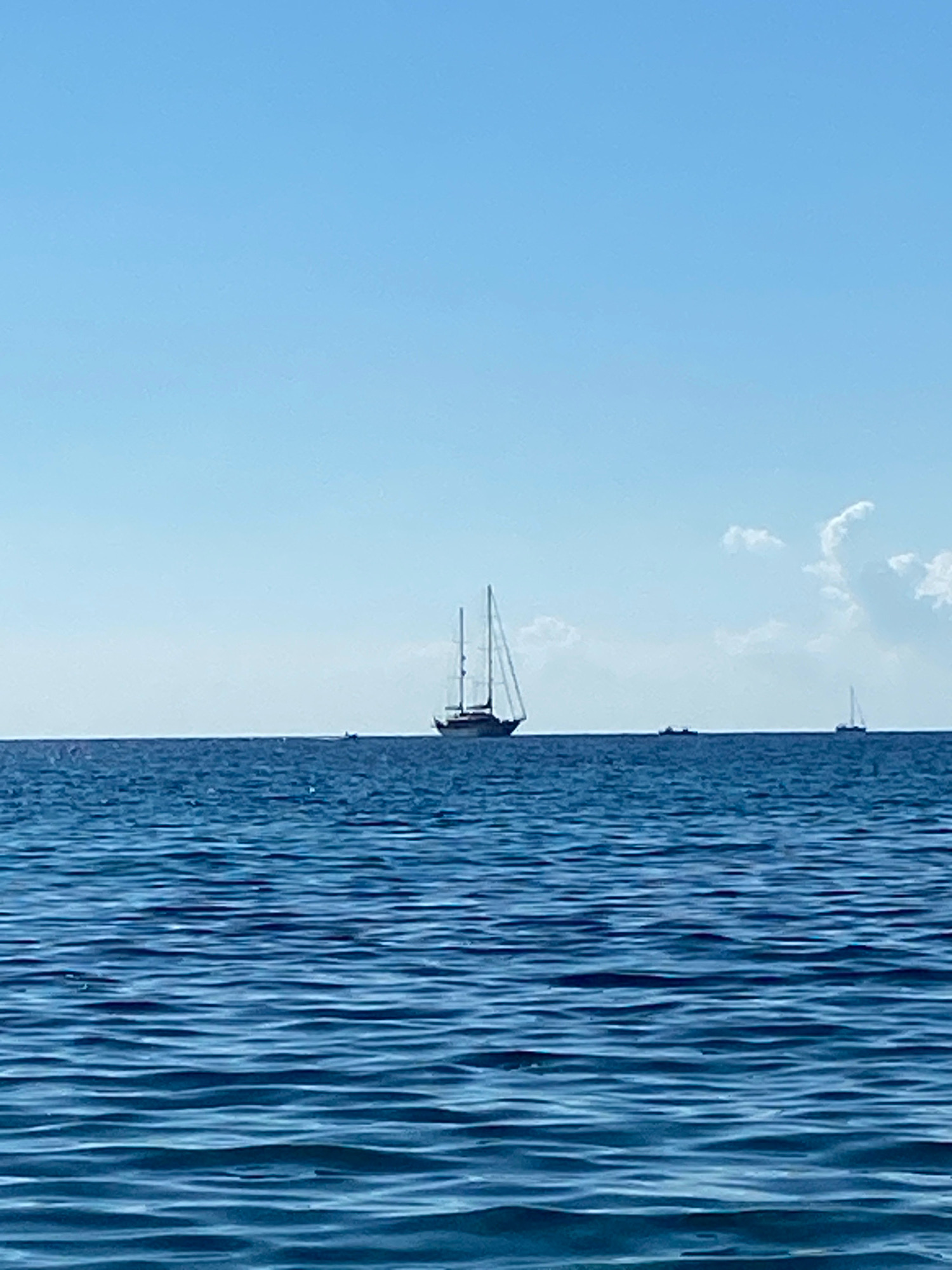 The image depicts a serene seascape under a clear blue sky with a few scattered white clouds. The ocean water is a deep blue, with gentle ripples creating a textured surface. On the horizon, several sailboats are visible. The most prominent is a large sailboat with its sails partially raised, positioned in the center of the image. To its right, a smaller sailboat with its sail fully extended is further in the distance. In between these two, a few smaller boats are also present. The scene evokes a sense of tranquility and a peaceful day at sea.