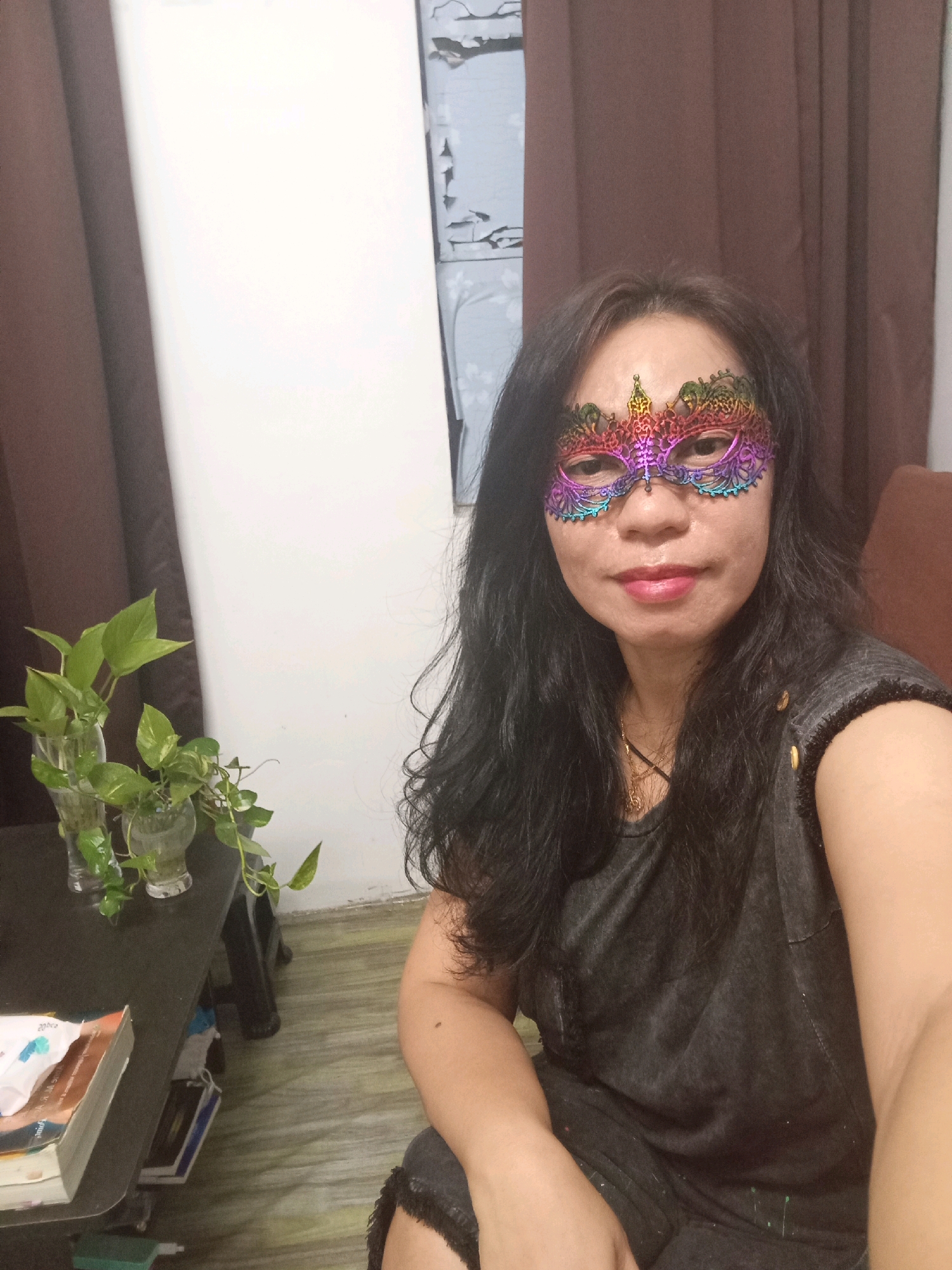 A medium shot captures a woman from the chest up, seated indoors in what appears to be a living room in Selayang Municipal Council, Malaysia. She has long, dark, wavy hair and is wearing a colorful, ornate mask that covers her eyes and the bridge of her nose, with a gold chain necklace visible underneath. Her top is a dark gray, sleeveless garment with a textured pattern and decorative buttons on the shoulder. She is looking directly at the camera with a neutral expression. To the left of the woman, a black table holds two glass vases with green plants. The background consists of brown curtains on the left and right, and a white wall in the center, with a partially visible patterned wallpaper or window covering. The lighting is dim, suggesting it is either evening or an overcast day.