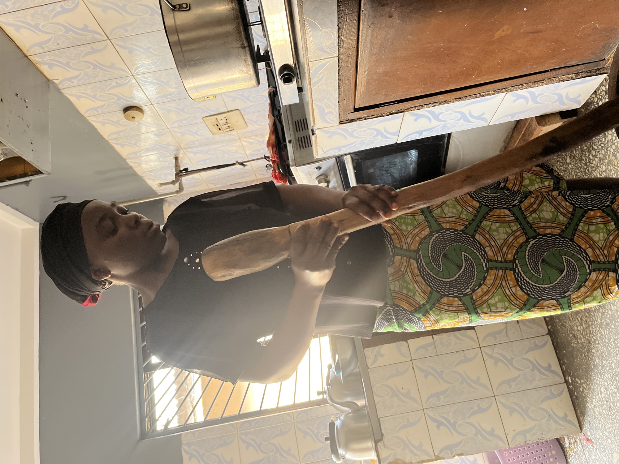 In a kitchen in Jos, Nigeria, a woman wearing a black headscarf and a patterned green and yellow skirt is holding a wooden object that looks like a pestle. The kitchen has tiled walls and floors. There is a stove and oven unit to her right and a sink to her left. Natural light is entering the room.