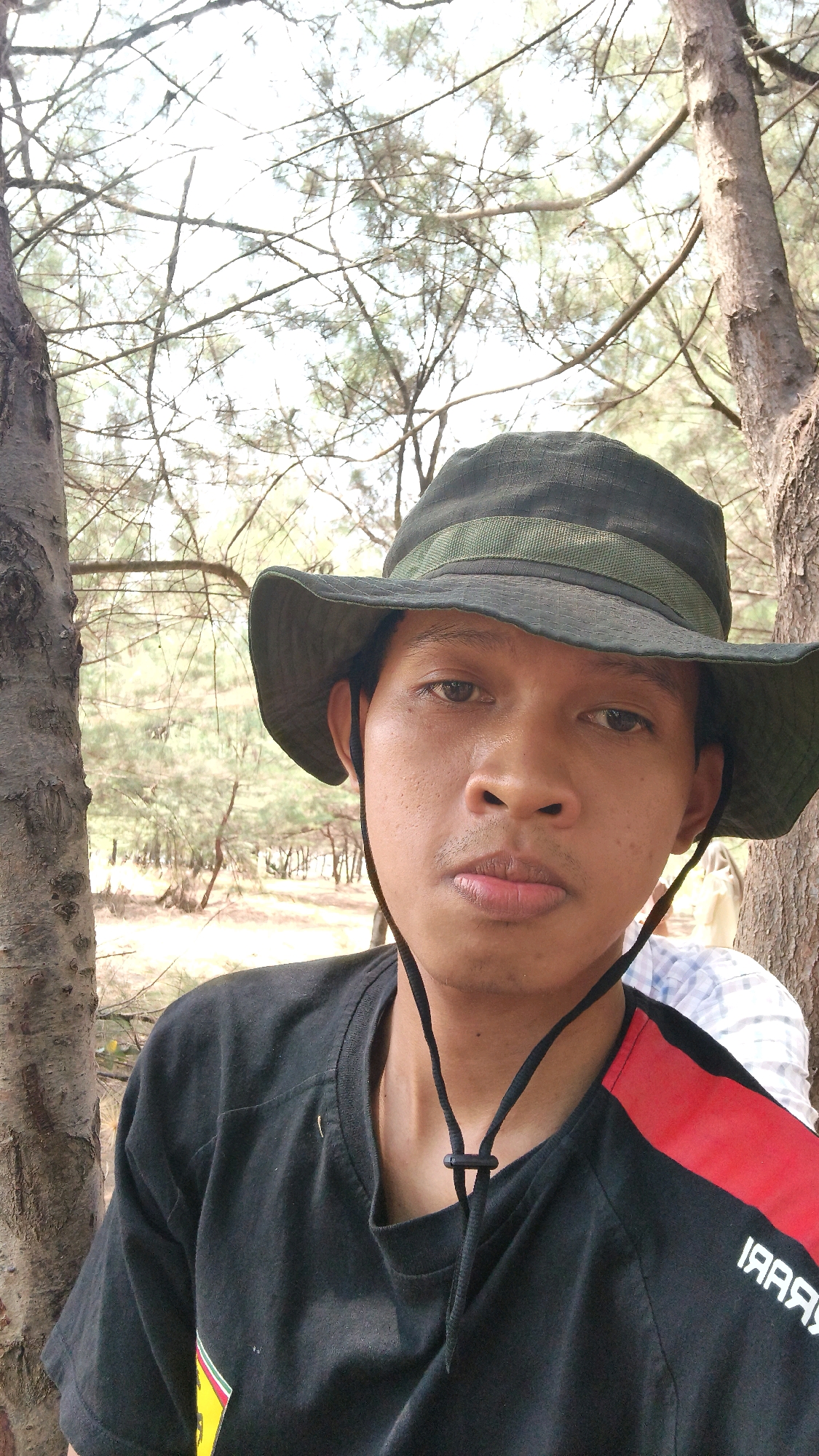 A person wearing a dark green wide-brimmed hat and a black t-shirt with a red and white diagonal stripe across the right shoulder is pictured in a selfie. The person's face is central in the frame, with the hat casting a shadow. The background consists of trees with sparse foliage and a bright sky, suggesting an outdoor environment. The person is positioned between two tree trunks, one on the left and one on the right, with the trunk on the right appearing to be a pine tree. The image appears to have been taken in Pabeanilir, Indonesia, given the presence of a beach area and coastal vegetation, typical of this Indonesian locale. The overall scene is casual and naturalistic.