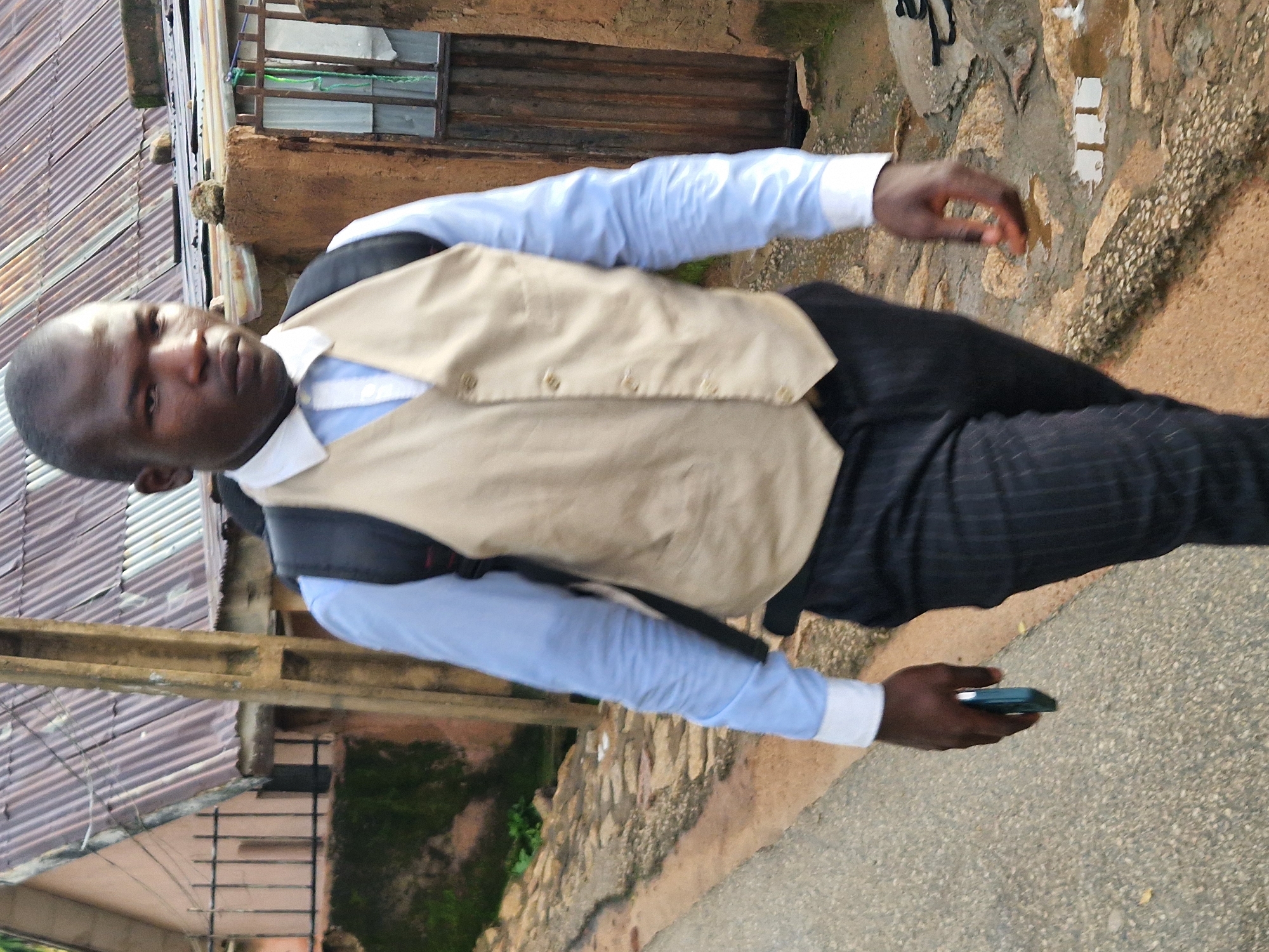 A man walks down a street in Kabong, Nigeria, carrying a backpack and a mobile phone. He is wearing a light blue dress shirt, a tan vest, and dark pinstripe pants. The street is paved with asphalt and bordered by a rocky embankment and a muddy ditch. A corrugated metal roof and a wooden door are visible in the background. The man is looking forward, his expression neutral.