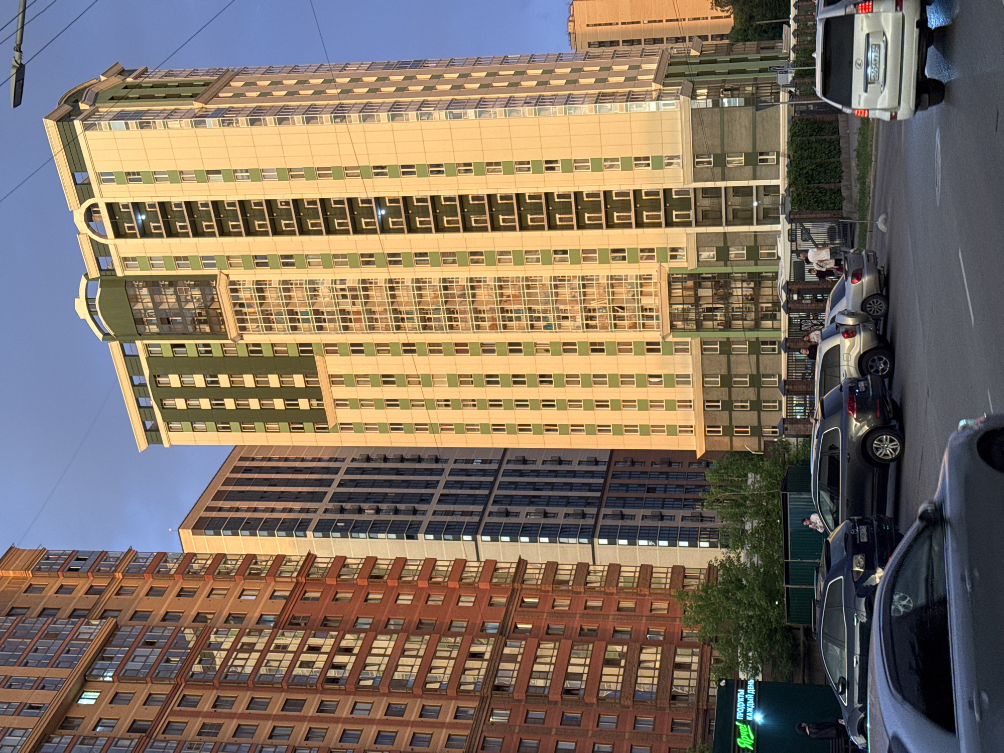The image captures a street view in Novosibirsk, Russia, featuring several tall residential buildings under a clear blue sky. The building on the right is a prominent, modern high-rise with a cream and green facade, featuring numerous windows and balconies. To its left, another tall building with a predominantly blue and white color scheme is visible. The leftmost building is a reddish-brown structure, also tall and with a facade punctuated by many windows. In the foreground, several cars are parked and driving on a paved street. Some trees and greenery are present between the buildings and on the ground level. A few people are visible on the sidewalks and near the cars. The lighting suggests it might be late afternoon or early evening, with the sun casting a warm glow on the buildings. A sign with Russian text is visible on the lower left, indicating a store or business. The overall scene depicts an urban residential area.