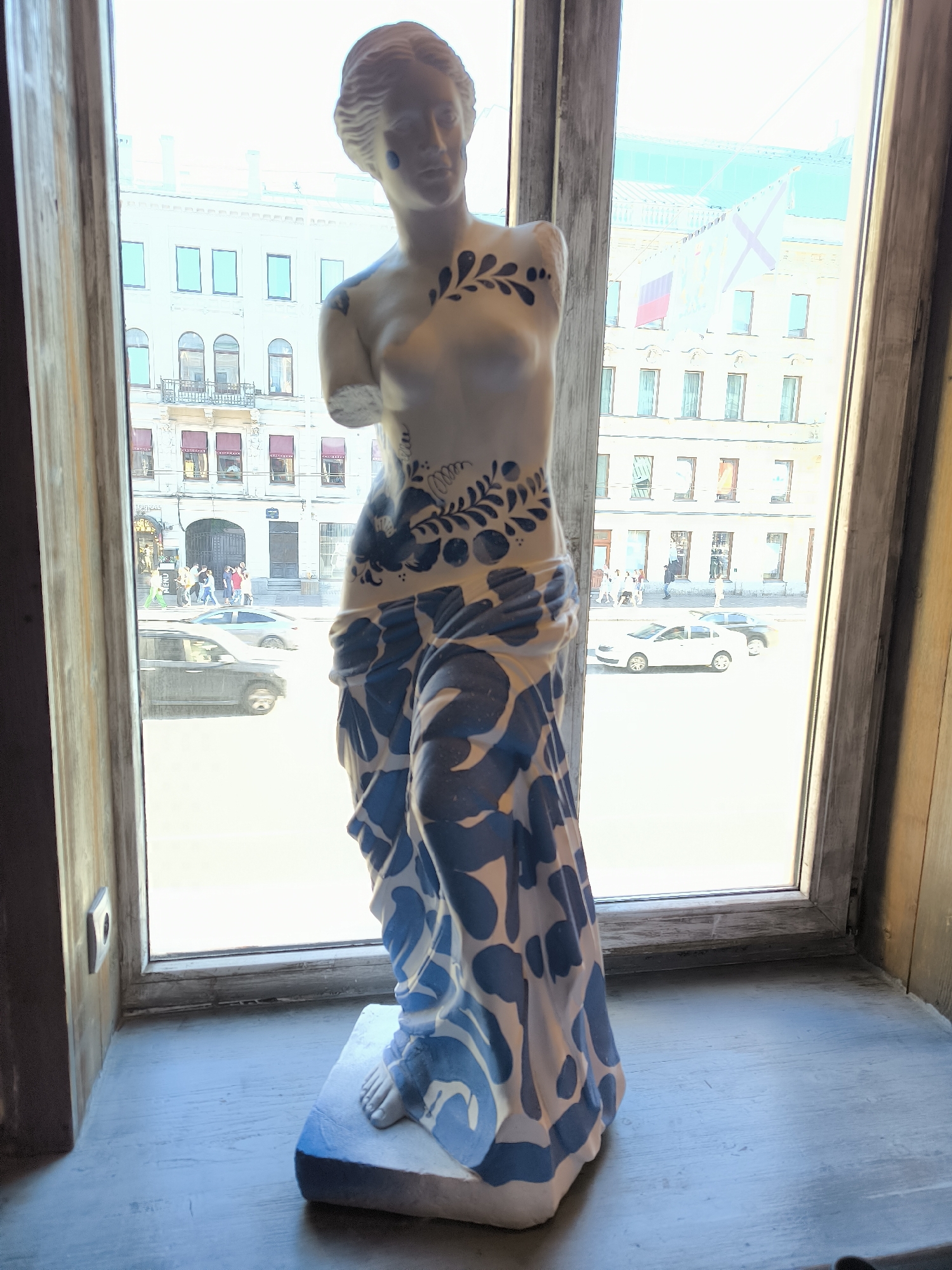 The image depicts a white, armless, classical-style female statue, approximately 1.5 meters tall, positioned on a grey-blue painted wooden window sill. The statue is decorated with intricate blue, Gzhel-style floral and abstract patterns across its torso and draped lower body, with two small blue teardrop motifs beneath its left eye. The statue's right foot is visible on a white base.
The statue is situated indoors, facing outward through a multi-pane window with distressed, light-grey wooden frames. Darker wooden paneling flanks the window recess.
Through the window, a brightly lit street scene in a city is visible. Opposite are multi-storied, light-colored (cream/pale yellow) buildings featuring a mix of rectangular and arched windows. Some windows have dark frames, while others are equipped with red retractable awnings or shades. Balconies with iron railings are also visible on some facades.
A building on the right side of the frame displays two prominent flags: a solid red rectangular flag and a white flag adorned with a blue saltire (St. Andrew's cross), which is the Russian Navy Ensign. A decorative banner is also partially visible near the flags.
At street level, pedestrians are visible walking on the sidewalks. Several vehicles are present on the street: a dark SUV in the foreground on the left, a silver sedan behind it, and a white sedan and a dark sedan further down the street on the right.
The presence of the Russian Navy Ensign (Andreevsky flag) and the distinct classical architectural style strongly indicate the location is in Saint Petersburg, Russia. The scene is illuminated by bright daylight, suggesting a sunny day, with the exterior view appearing somewhat overexposed compared to the interior.