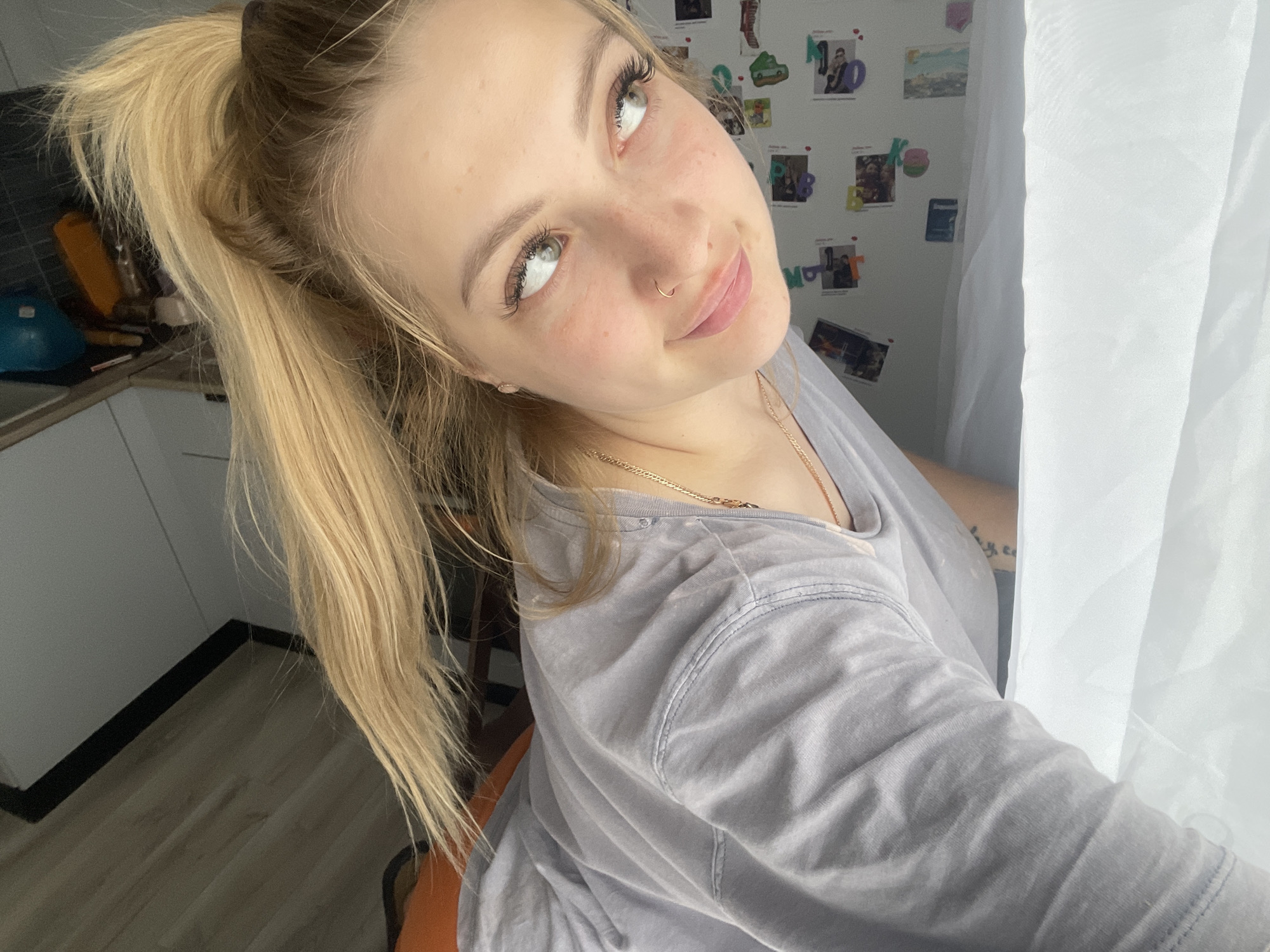 A young woman with blonde hair pulled back in a ponytail is positioned in what appears to be an indoor setting, possibly a home. She is wearing a light gray t-shirt. She has a nose ring and is wearing a gold necklace. The woman is looking upwards and to the side with a slight smile. Behind her, there is a wall decorated with photographs and letter magnets. To the left, a kitchen area is visible. Next to her is a white curtain.
I am unable to provide information about location in the city of Izhevsk, Russia.