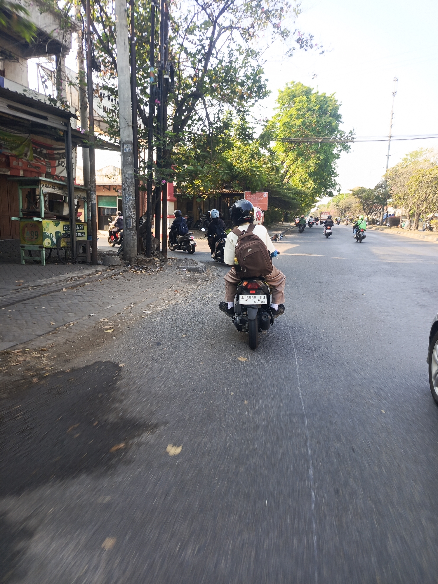 A street scene in Ngampelsari, Indonesia, features a light pole on the left, with buildings and trees lining the road. Several people are riding motorcycles, most of whom are wearing helmets. In the foreground, a person wearing a helmet and a backpack is riding a motorcycle. The motorcycle has a license plate that reads "W-6589-UJ". The road appears to be paved, with some debris on the side. The sky is clear and blue, indicating a sunny day.