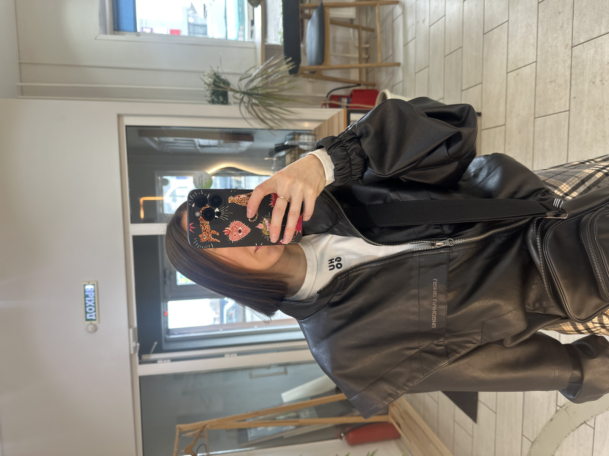 A woman is taking a selfie indoors. The woman is wearing a white shirt and a black leather jacket. The phone has a decorative floral case.
It is not possible to confirm the location is Votkinsk, Russia.