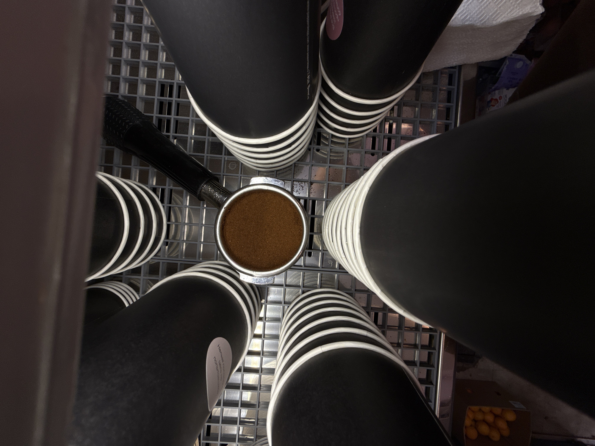 The image shows a close-up, overhead view of a coffee-making setup. The main subjects are several black disposable cups with white stripes, stacked and arranged in a radial pattern around a portafilter filled with ground coffee. A black tamper is visible resting against the portafilter. The cups appear to be made of paper. The portafilter and coffee grounds are centered in the frame.
The setting appears to be a commercial kitchen or coffee preparation area, with a metal grid surface beneath the cups. In the background, a white towel or cloth is partially visible, along with what looks like a small cardboard box containing yellow objects, possibly lemons, and a purple box with some blue and red graphics, which could be a carton of milk or juice. The overall lighting suggests an indoor environment, possibly with artificial light, though the specific time of day is not discernible.
There is some faint, light-colored text on a sticker on one of the cups, but it is not legible. The scene is static, focusing on the preparation of coffee, with no people or explicit activity other than the objects themselves. The context is provided as Dolgoprudny, Russia.