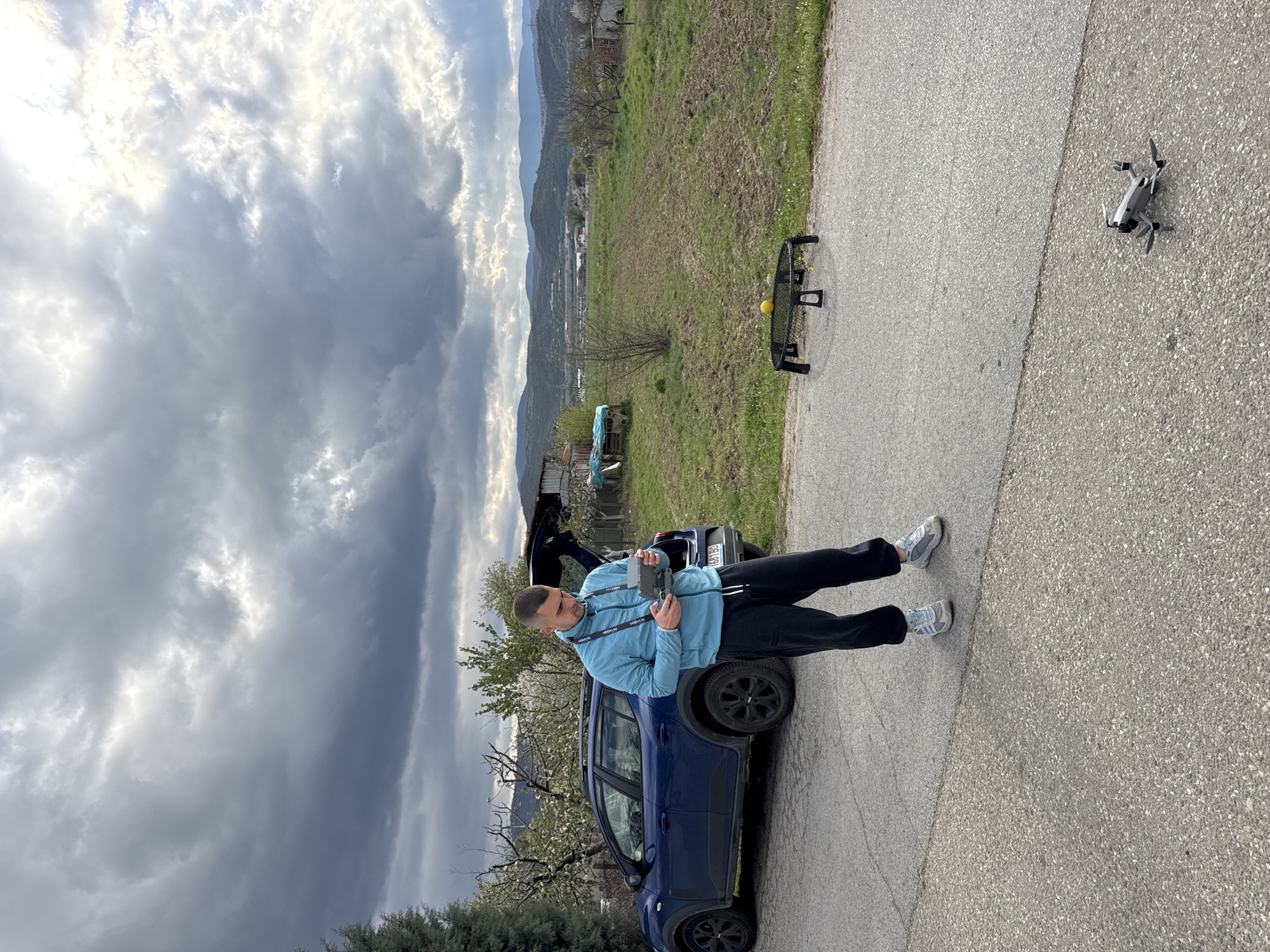 A slightly high-angle, outdoor shot shows a man standing on a paved road, holding a controller, presumably for the white drone hovering nearby.
The man is light-skinned with short dark hair, wearing a light blue jacket, black pants, and grey and white sneakers. He is standing next to a dark blue car with the trunk open. Behind the car is a partially obscured view of a grassy field.
To the left, the sky is overcast with heavy clouds. In the background, there are rolling hills and a glimpse of a town or city. There is a black net and a yellow ball on the right-hand side of the background.