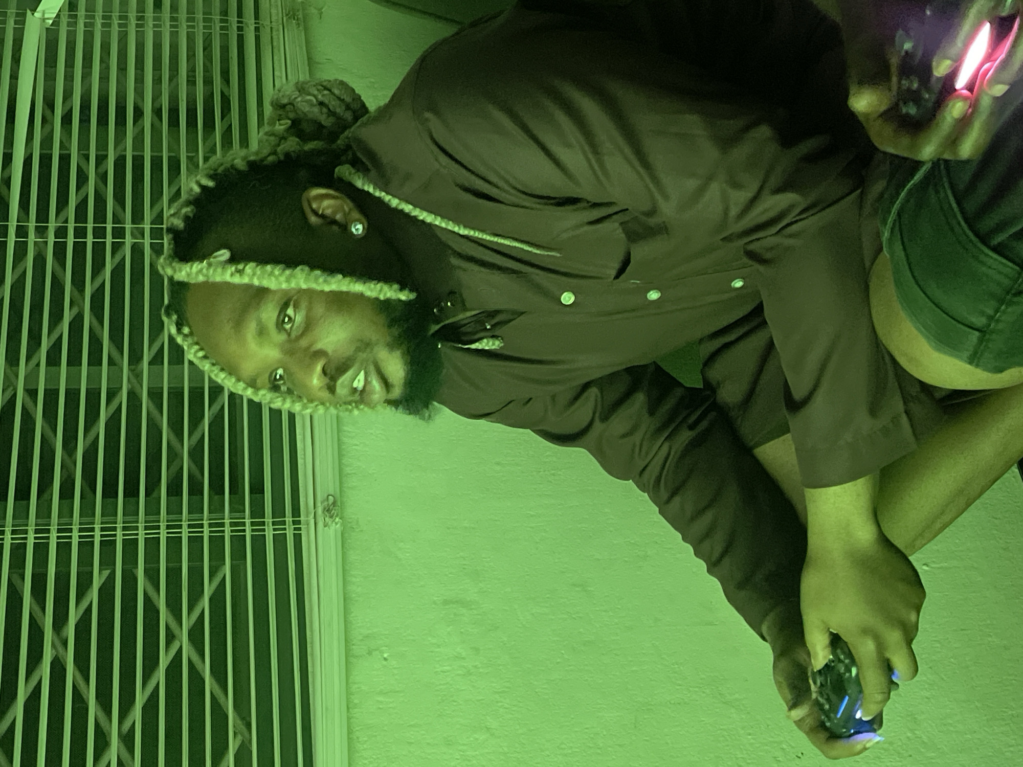 In a dimly lit room in Jos, Nigeria, illuminated by green lighting, a man with blonde-streaked braids is intensely focused on playing a video game. He is seated, wearing a dark button-up shirt and shorts, with a gaming controller in his hands. The background features a wall and horizontal blinds. The green lighting casts a distinct hue over the entire scene, highlighting the textures of the wall and the man's skin.