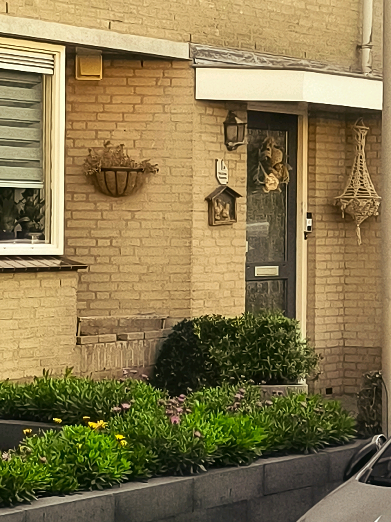 The image depicts a close-up view of the exterior of a brick house, likely in the city of Oostvoorne, Netherlands. The house features a window with horizontal blinds on the left and a dark-colored front door on the right. Above the door, a lantern is mounted to the brick wall. A decorative wreath hangs on the door. To the right of the door, a macrame-style hanging decoration is suspended from the wall. In front of the house, a garden bed is filled with lush green foliage and a scattering of small pink and yellow flowers. The garden bed is bordered by a dark grey stone planter. A portion of a car is visible in the lower right corner of the frame. The lighting suggests it is either early morning or late afternoon.