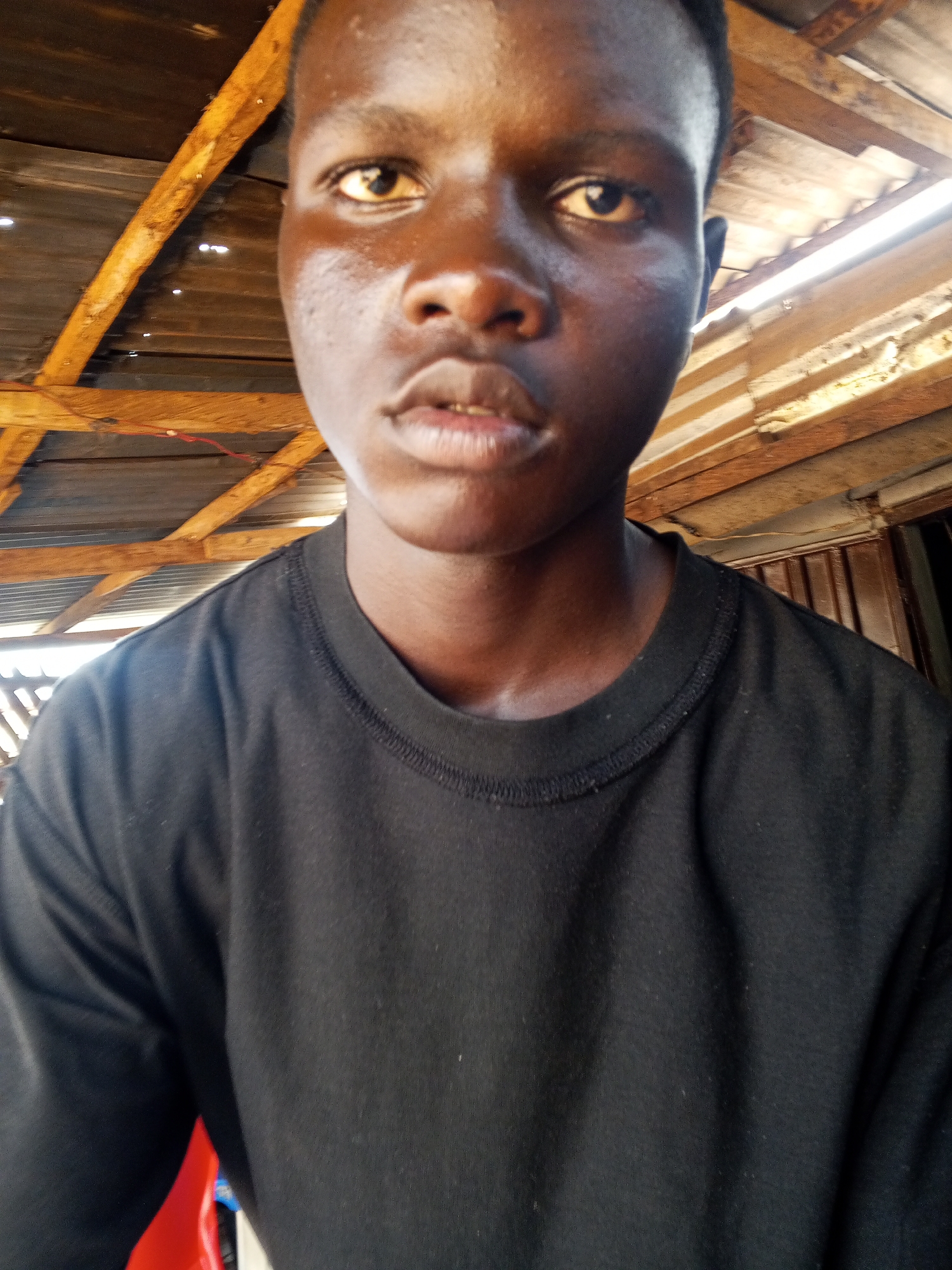 A young male with dark skin and short dark hair is depicted in a head-and-torso portrait, directly facing the camera. He wears a plain black long-sleeved t-shirt with a ribbed crew neck. His eyes appear light brown or amber due to the bright light source, and his lips are slightly parted. The background reveals an interior structure featuring a corrugated metal ceiling supported by visible, rough-hewn wooden beams. Gaps in the ceiling allow bright, direct natural light to illuminate the scene, creating strong highlights and shadows. The overall environment appears to be a basic, possibly unfinished or rustic indoor space. The scene is situated in the city of Unknown, Unknown.