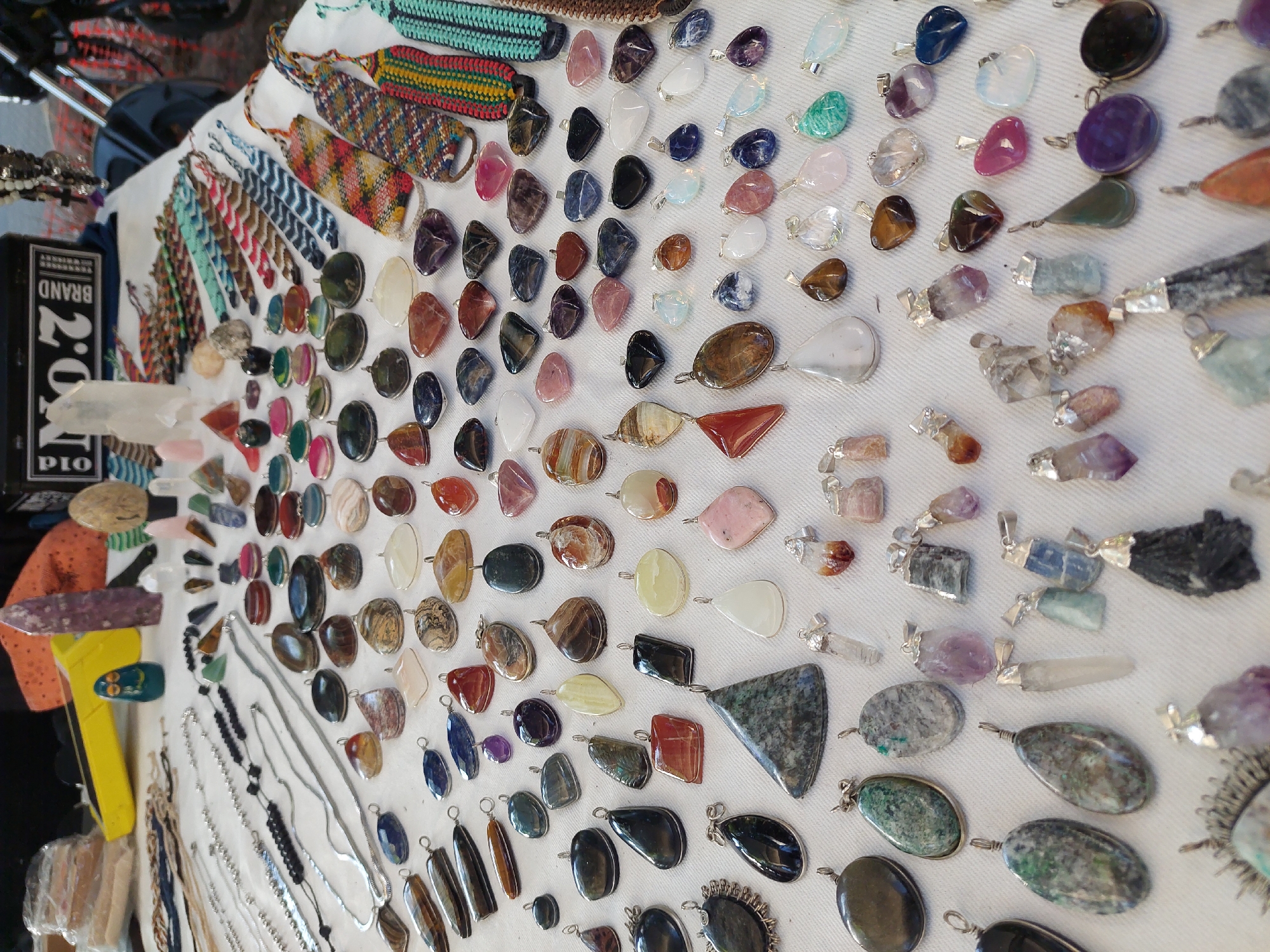 A high-angle, slightly skewed view shows a display of jewelry and woven bracelets arranged on a white fabric surface. The jewelry consists of numerous pendants made from various gemstones and minerals, each attached to a silver or metal finding for stringing onto a necklace. The pendants vary in shape, size, and color, ranging from polished stones to rough crystal formations. Adjacent to the pendants are several woven bracelets in a variety of colorful patterns. Near the left edge of the image, various silver chains and additional jewelry are also visible. A black box with white lettering sits near the bracelets. The scene suggests a marketplace or craft fair where these items are being sold. Given the presence of handcrafted items and the bustling atmosphere often associated with artisan markets, the location could be a street fair or artisan market in Buenos Aires, Argentina.