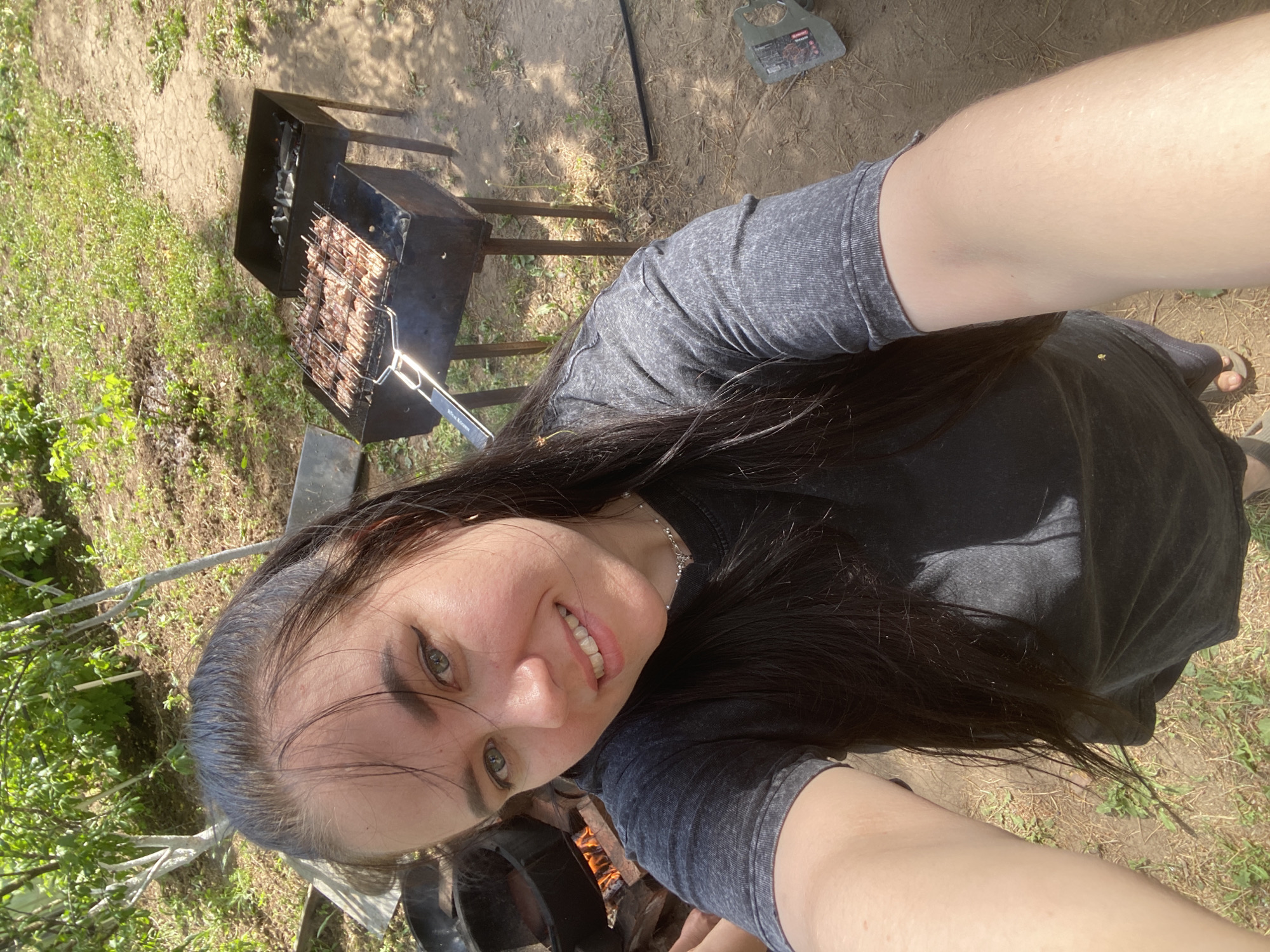 A selfie shows a woman with long, dark hair, wearing a gray t-shirt, smiling at the camera. Behind her, a barbecue grill is positioned over a bed of burning coals, with food grilling on the grate. The setting appears to be an outdoor area with grass and dirt. A garden tool sits on the ground next to the grill. The image's location is Chaikovsky, Russia.
