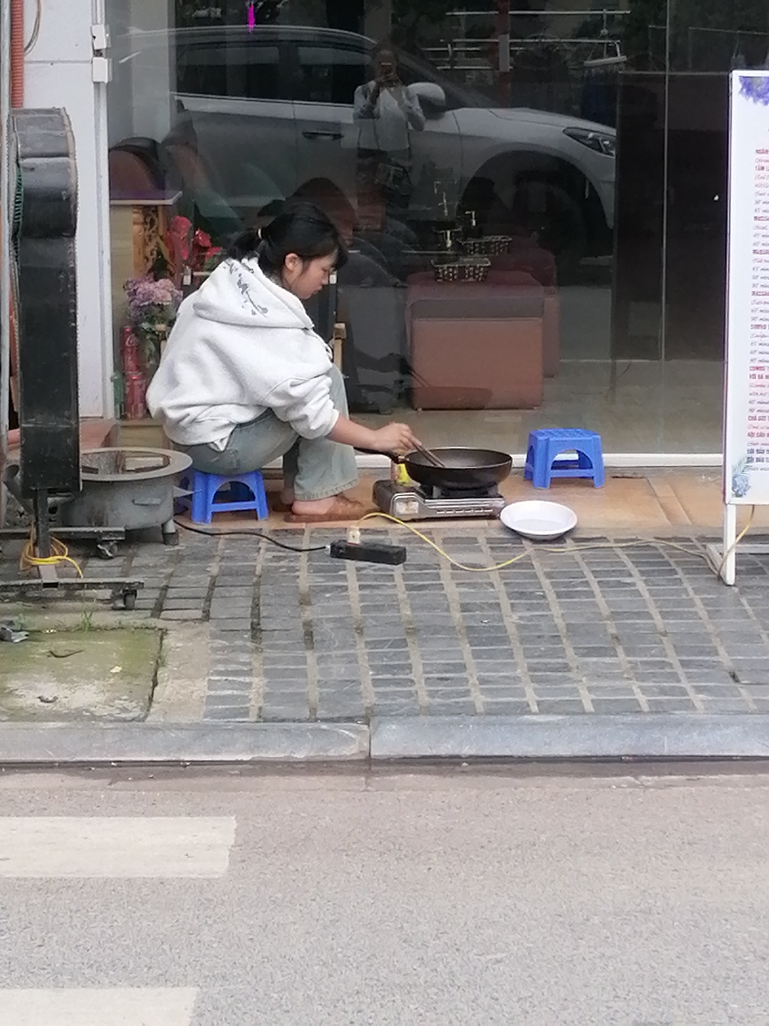 A young woman with dark hair pulled back in a ponytail is squatting on a small blue plastic stool and cooking in a wok with chopsticks. She is wearing a white hooded sweatshirt and blue jeans, and brown sandals. The cooking is taking place on a portable gas stove. A white bowl sits to the right of the stove and wok. Behind her is a window with reflections of buildings and a white car. To her left is a black metal stove with yellow wires, and a menu board is to her right. The scene is taking place on a sidewalk made of gray bricks. The image location is identified as Sa Pa Ward, Vietnam.