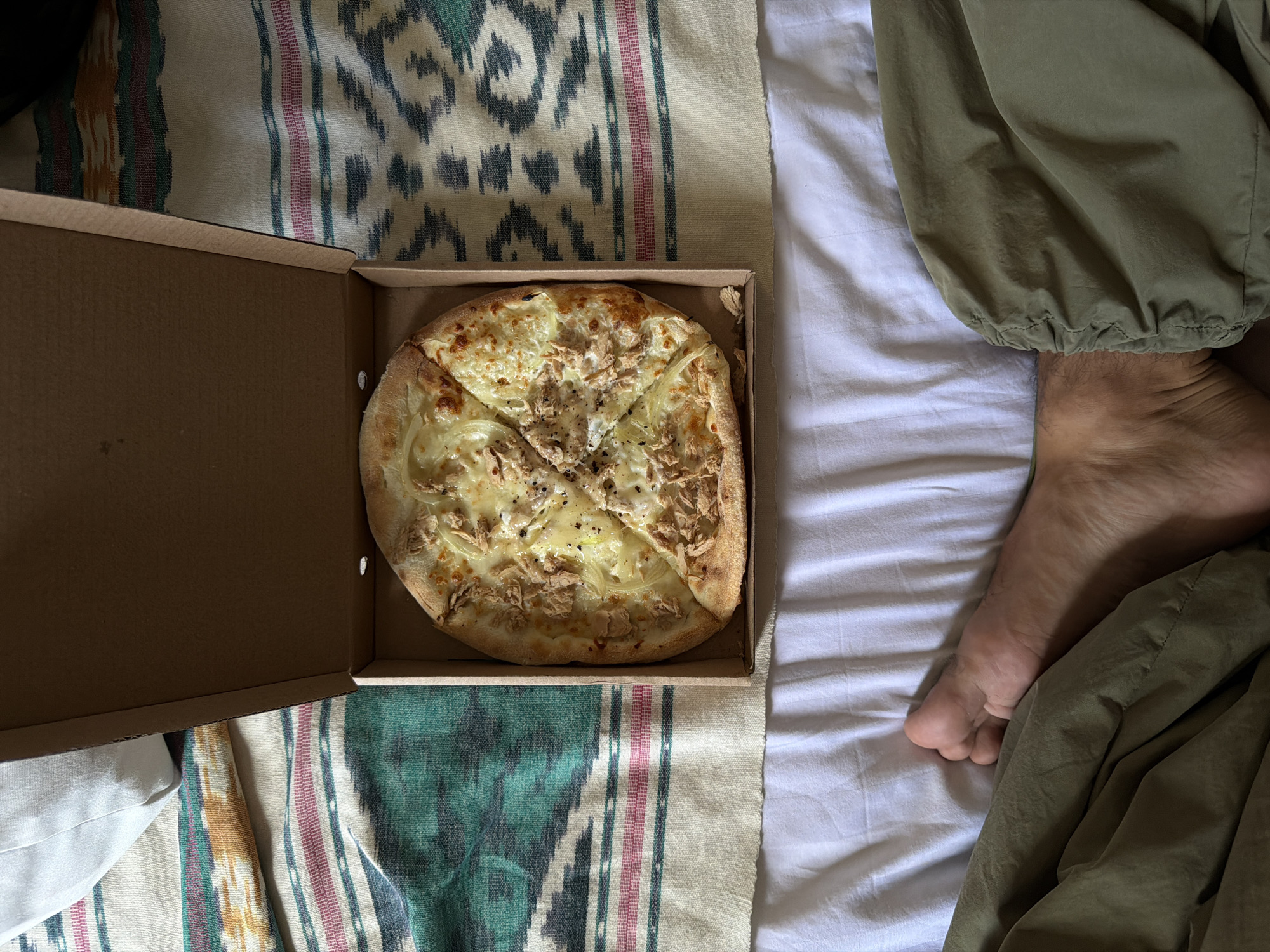 A medium-sized pizza, cut into four slices, sits in a cardboard box on a multi-colored fabric. White sheets and a person's foot are visible next to the pizza box, partially obscured by green clothing. The location is unspecified, but based on metadata, it is in North Kuta, Indonesia.