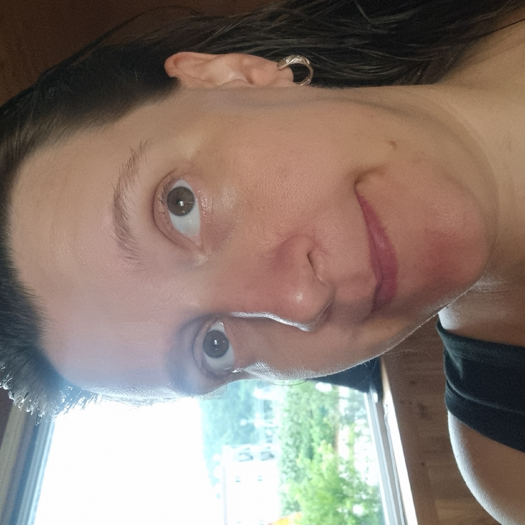 The image shows a close-up, waist-up, outdoor selfie of a woman with dark hair and brown eyes, wearing a black tank top. She has a hoop earring in her right ear and appears to be looking slightly upwards. A window with a view of greenery and buildings is visible in the background. The woman is positioned indoors, with the window behind her. The context of Dunnae-myeon, South Korea, is not visually evident in the image.