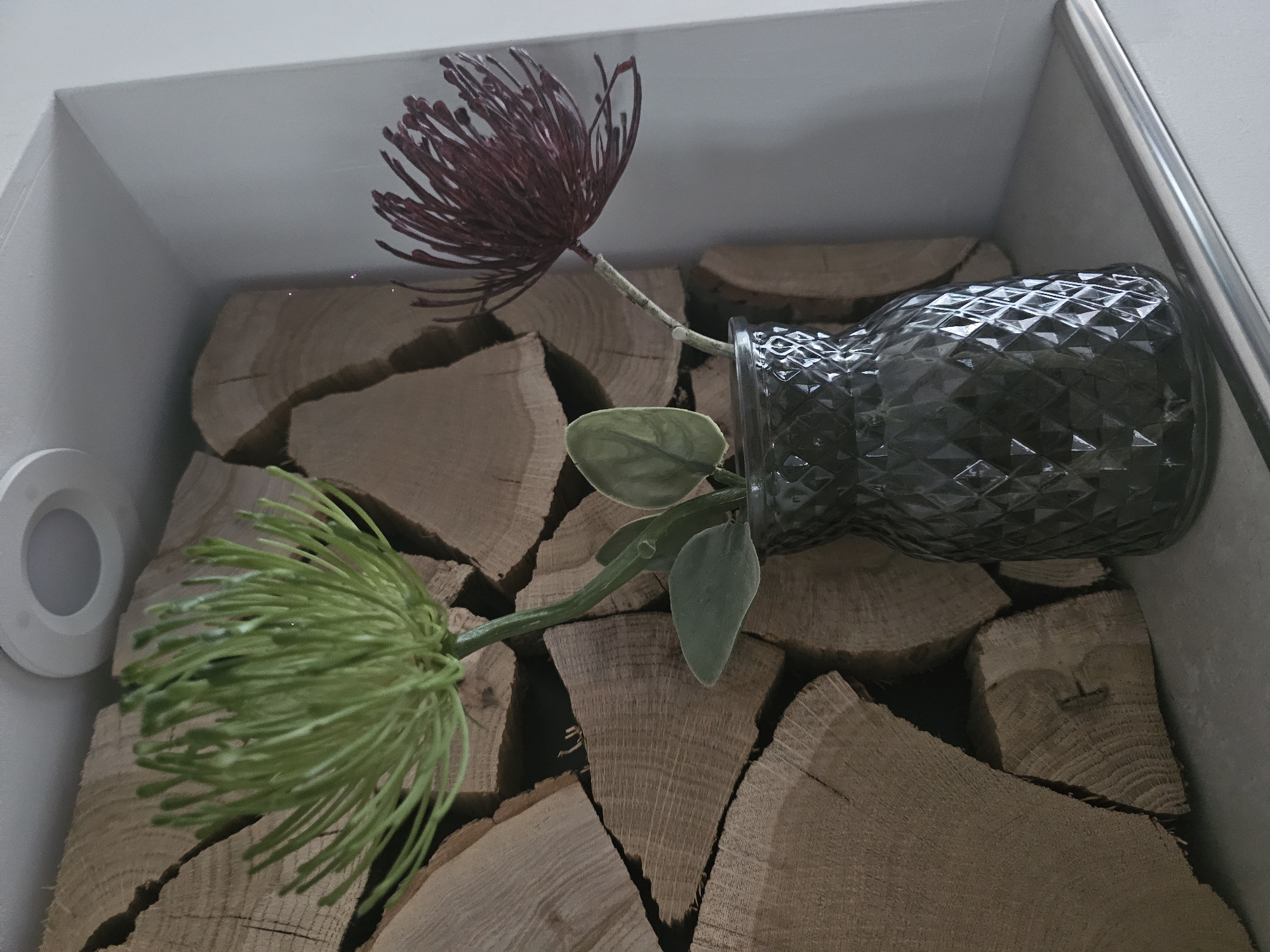 An interior scene shows two artificial flowers in a dark, patterned glass vase. The vase sits on a gray surface, and behind it is a wall of stacked logs. A ceiling light fixture is visible in the upper portion of the image. One flower is bright green and spiky, while the other is a deep red-purple and has a similar spiky texture. The logs are rough-hewn and have visible growth rings. The scene is lit from above, casting shadows on the logs.