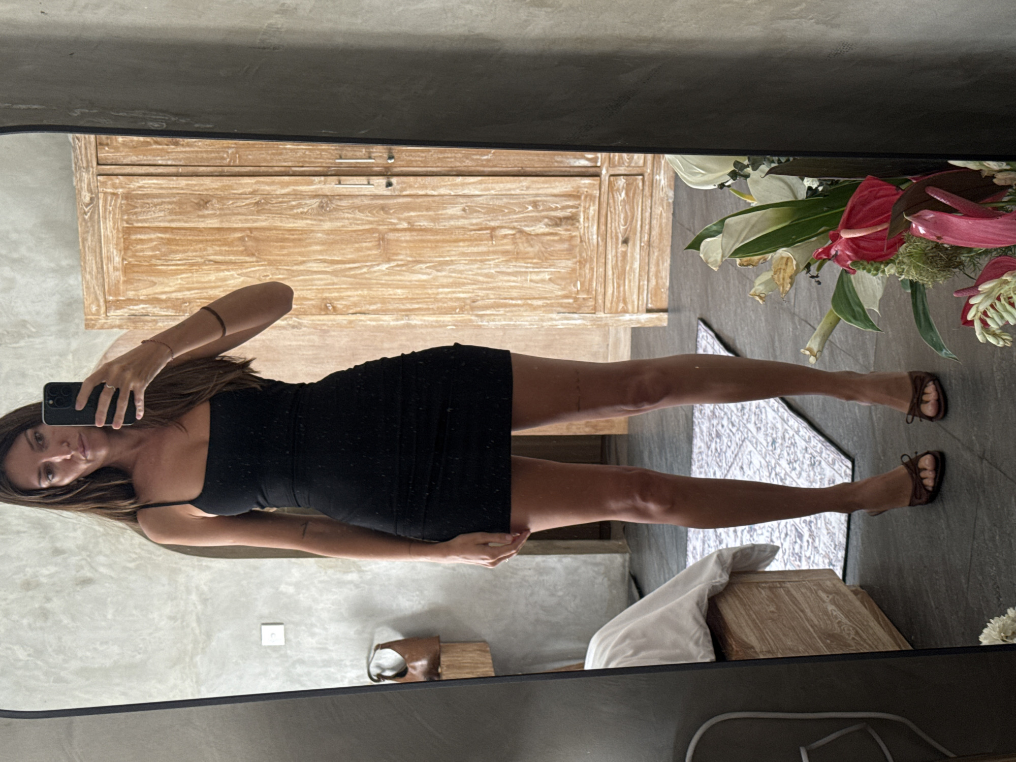 A woman is reflected in a mirror, holding a smartphone, presumably taking a selfie. She wears a black, sleeveless dress with thin straps and brown sandals. Behind her, there's a wooden cabinet with drawers and a floral arrangement. The background includes a wall with a concrete-like texture, suggesting an indoor setting. Based on the details and the user prompt, the location is likely a hotel or residence in North Kuta, Indonesia.
