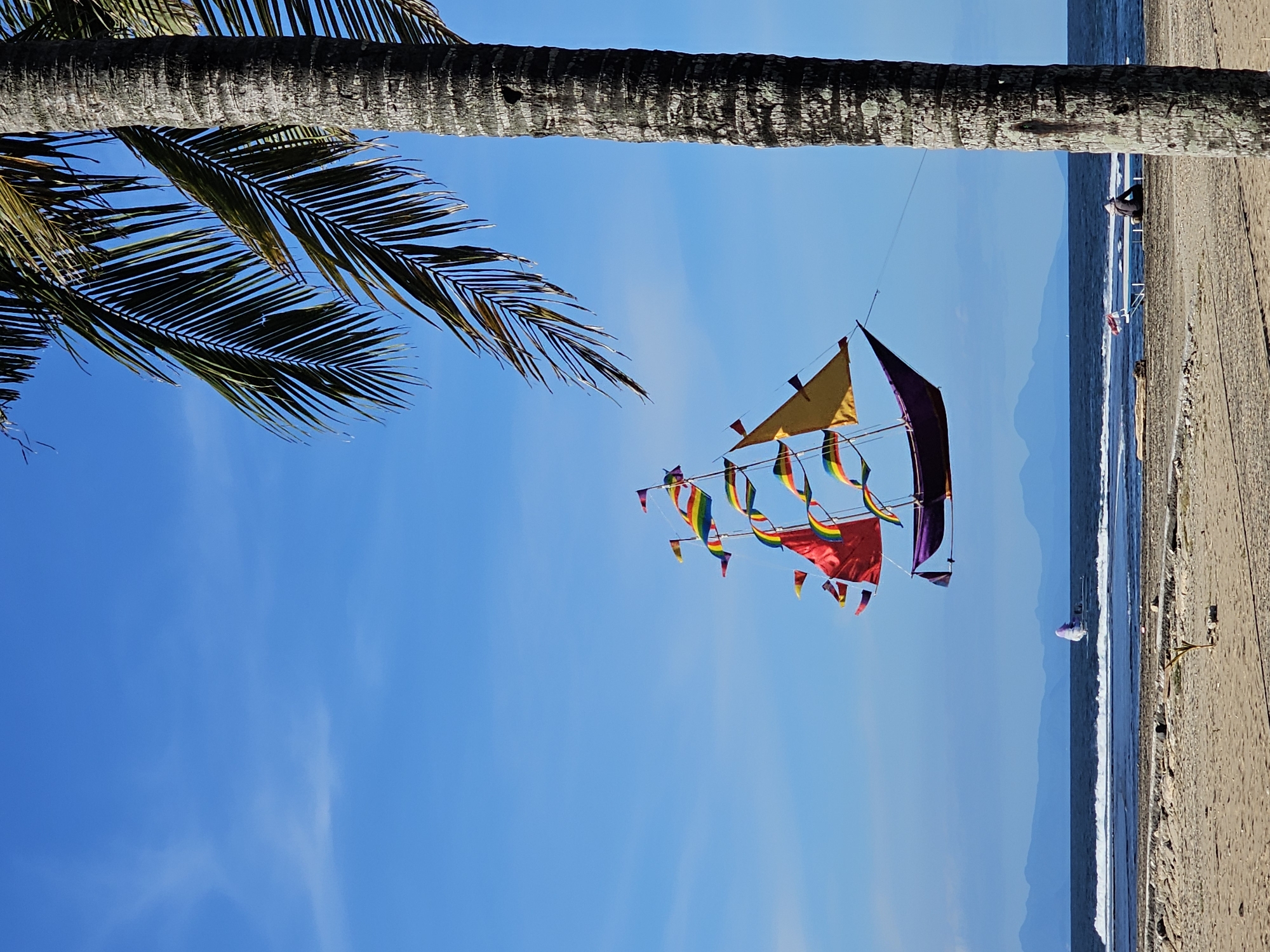 The image shows a colorful kite resembling a ship in the sky of Kuta Selatan, Indonesia. A palm tree trunk is on the upper left, and a beach with a person sitting is on the right. A mountain range is in the distance.