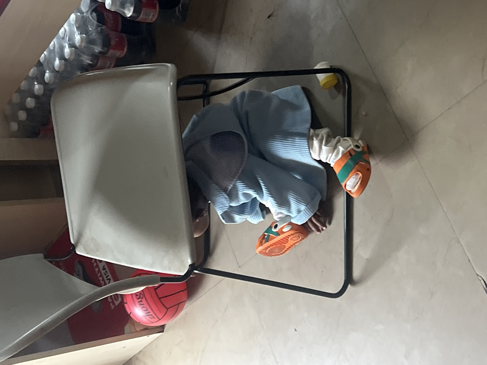 A child is positioned under a white chair, possibly hiding or playing. The child is wearing a light blue garment and orange shoes. The chair is placed on a tiled floor and has a black metal frame. A red volleyball is positioned beside the chair. Shelves containing stacked items, likely beverages in plastic packaging, are visible in the background. The location, Gura Top, Nigeria, cannot be determined from the image alone.