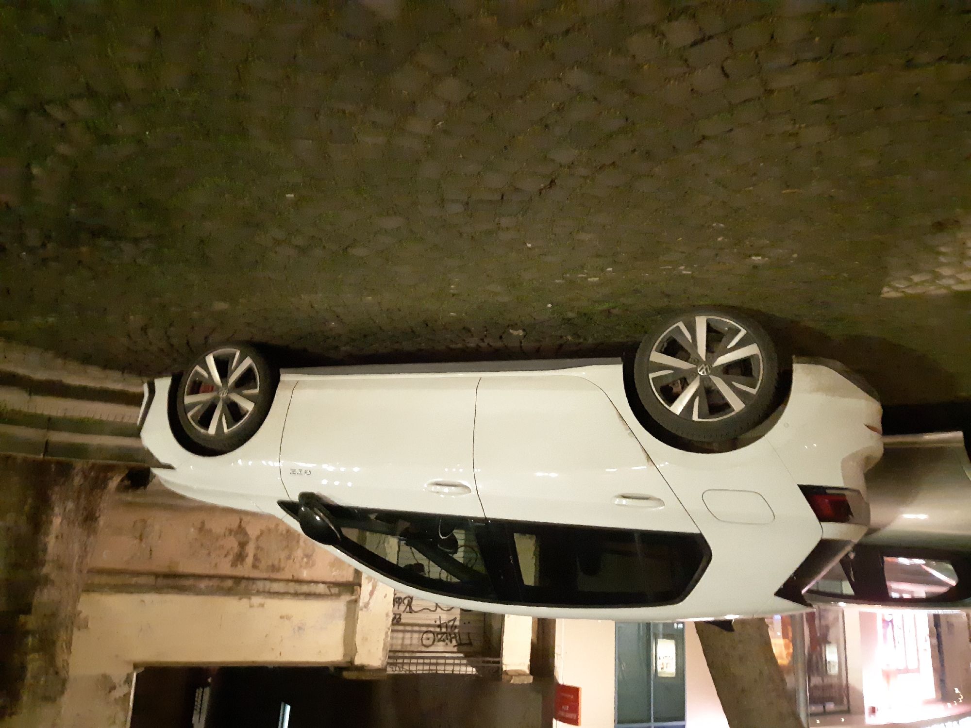 A white Volkswagen Golf GTE is parked on a cobblestone street in Plovdiv, Bulgaria. The car is oriented with its front facing right. The vehicle's wheels and body are clearly visible. The surrounding environment includes buildings with doors and windows, as well as a wall with graffiti. The ground is paved with irregular stones or cobblestones. The lighting suggests it's nighttime. There are no people visible in the scene.