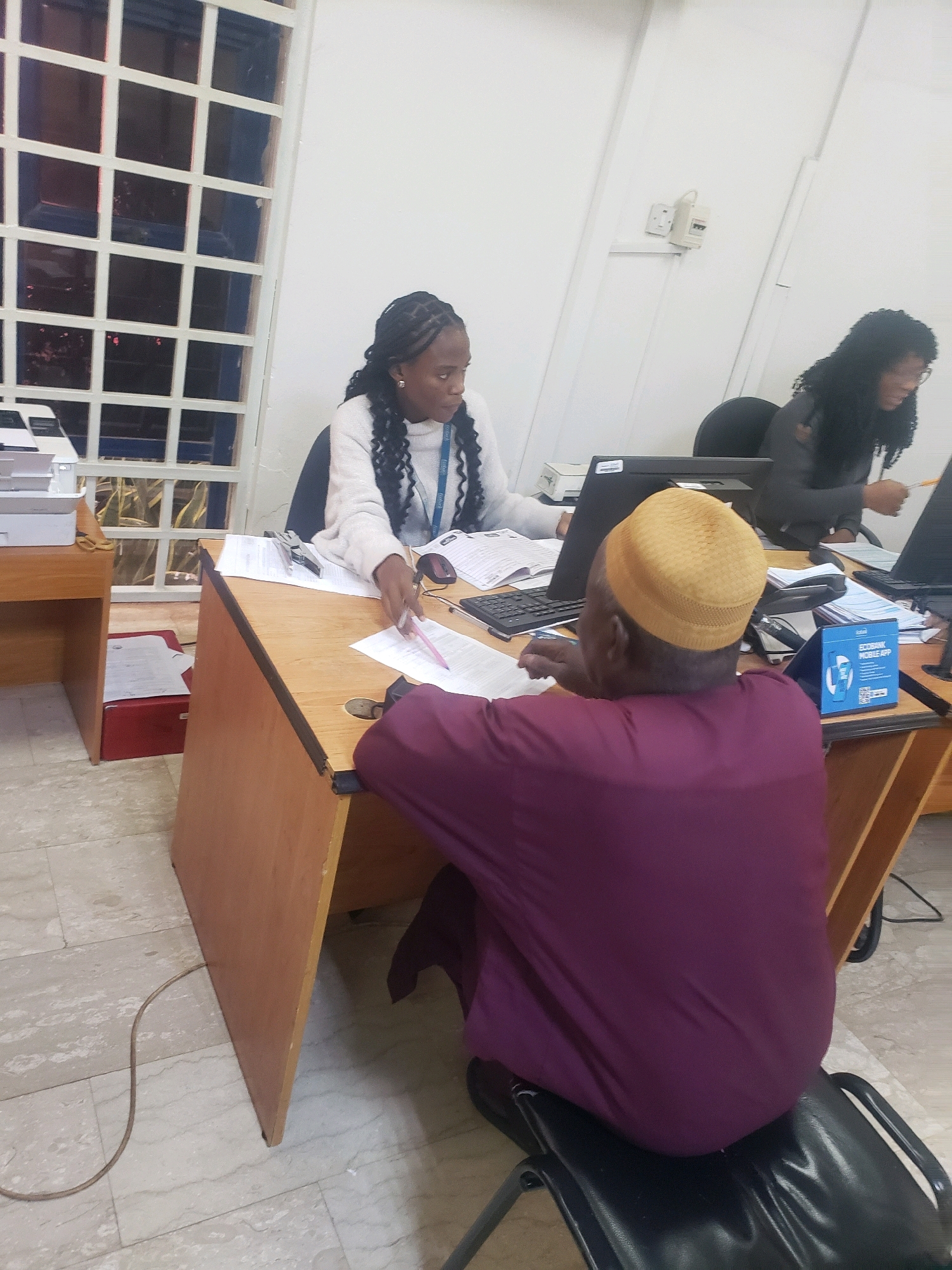 In Kakuri Gwari, Nigeria, two women and one man are engaged in administrative work within an office setting. The scene depicts a woman with braided hair wearing a light-colored sweater, seated at a wooden desk. She is actively working, with papers and a pen in hand. To her right, another woman, possibly a colleague, is also seated at a desk, her attention focused on a computer monitor. In the foreground, a man wearing a maroon robe and a yellow cap is seated and appears to be reviewing documents. The office environment includes desks, computers, telephones, and a prominent sign for the "ECOBANK MOBILE APP". A tiled floor and a white wall with electrical outlets complete the indoor setting.