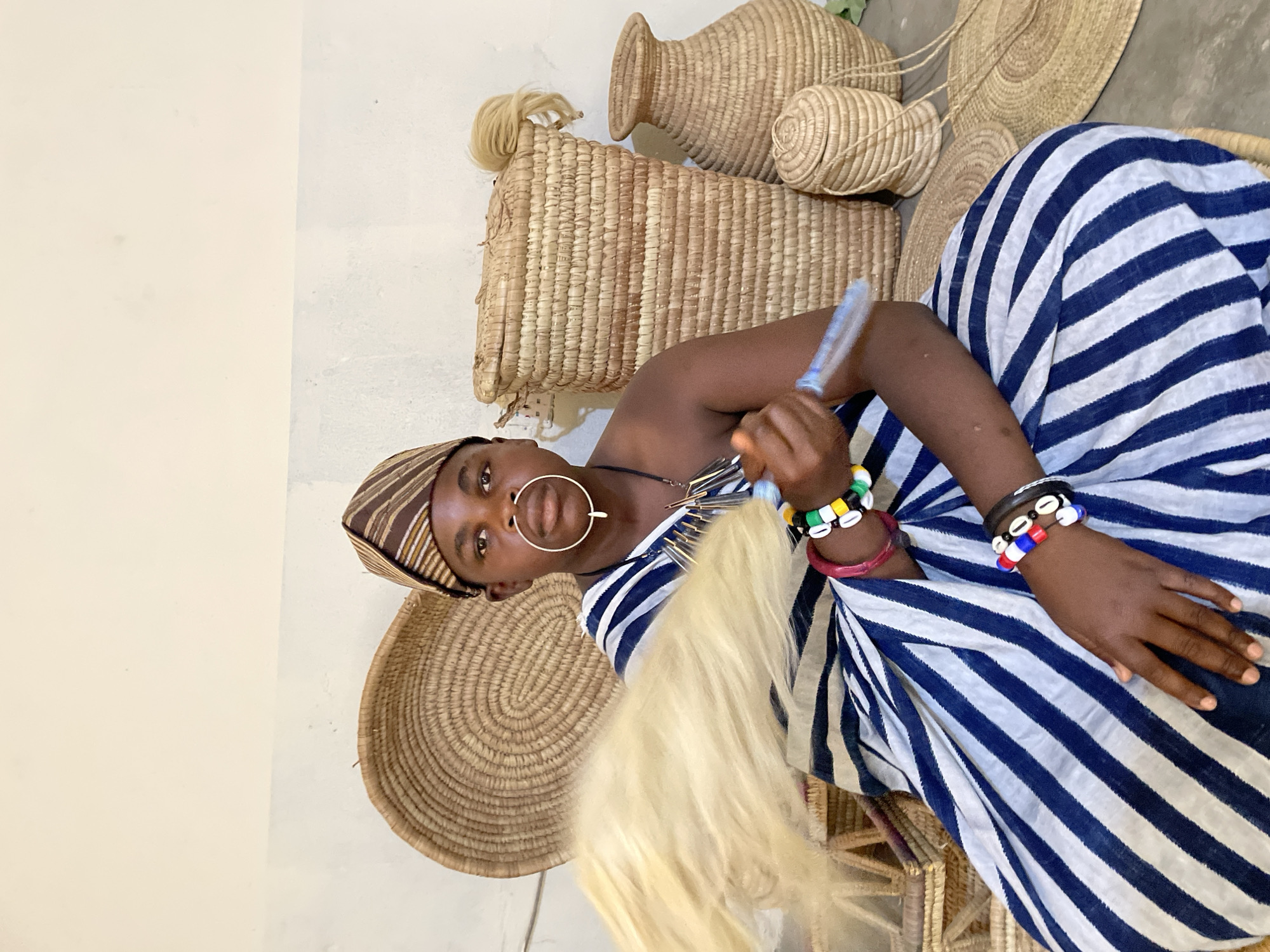 An individual, likely a model, is positioned amidst a collection of woven baskets and hats, dressed in attire that suggests cultural or ceremonial significance. The person is wearing a striped garment with a headdress, a large nose ring, and bracelets featuring colorful beads and shells. One hand holds a bundle of fibers, possibly used for craft or ritualistic purposes. The location is specified to be in the city of Jos, Nigeria.