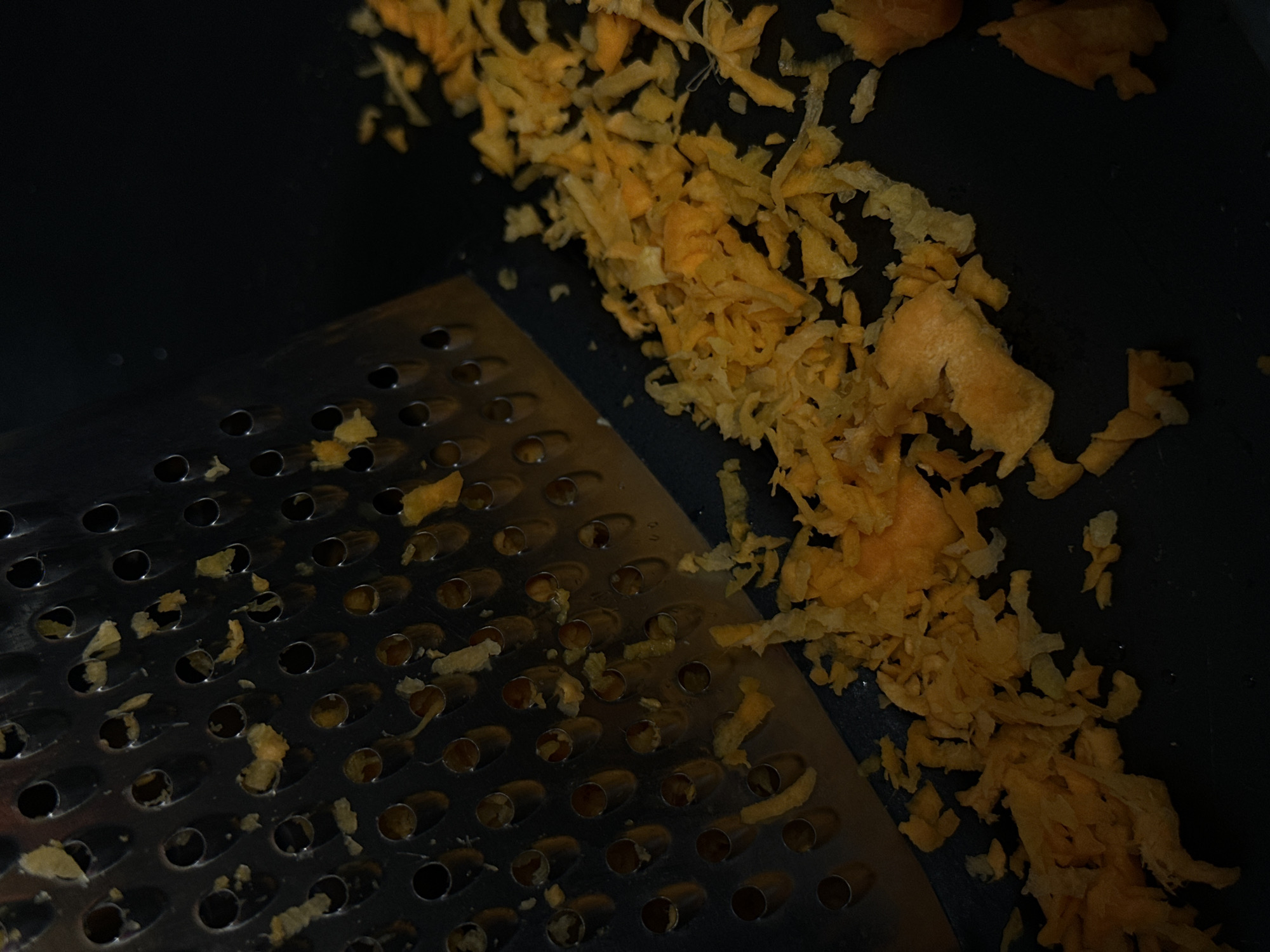 A close-up shot shows a grater resting on a black surface. Shredded orange material is scattered around the grater and on the black surface. The grater is metallic with numerous circular holes. The composition focuses on the grater and the shredded material, with a dark and neutral background. There's no information to suggest the image was taken in the city of Chaikovsky, Russia.