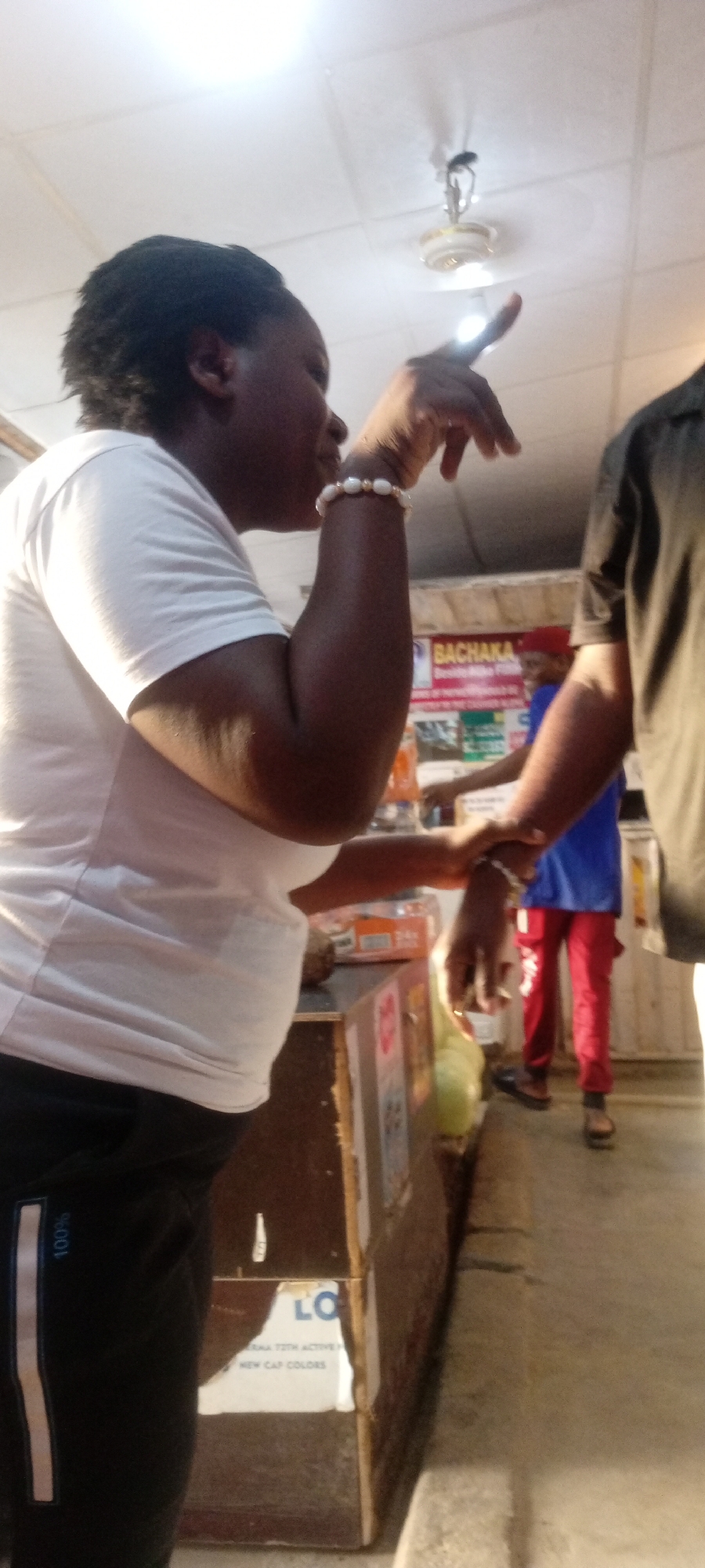 A person wearing a white t-shirt, black pants, and a beaded bracelet points towards a ceiling light. Another person is wearing a black shirt and standing beside the first person. In the background, a person wearing a blue shirt and red pants is walking. A counter with products sits in the foreground. A sign with "BACHAKA" is visible in the background. The location is presumed to be a retail establishment in Kano, Nigeria.
