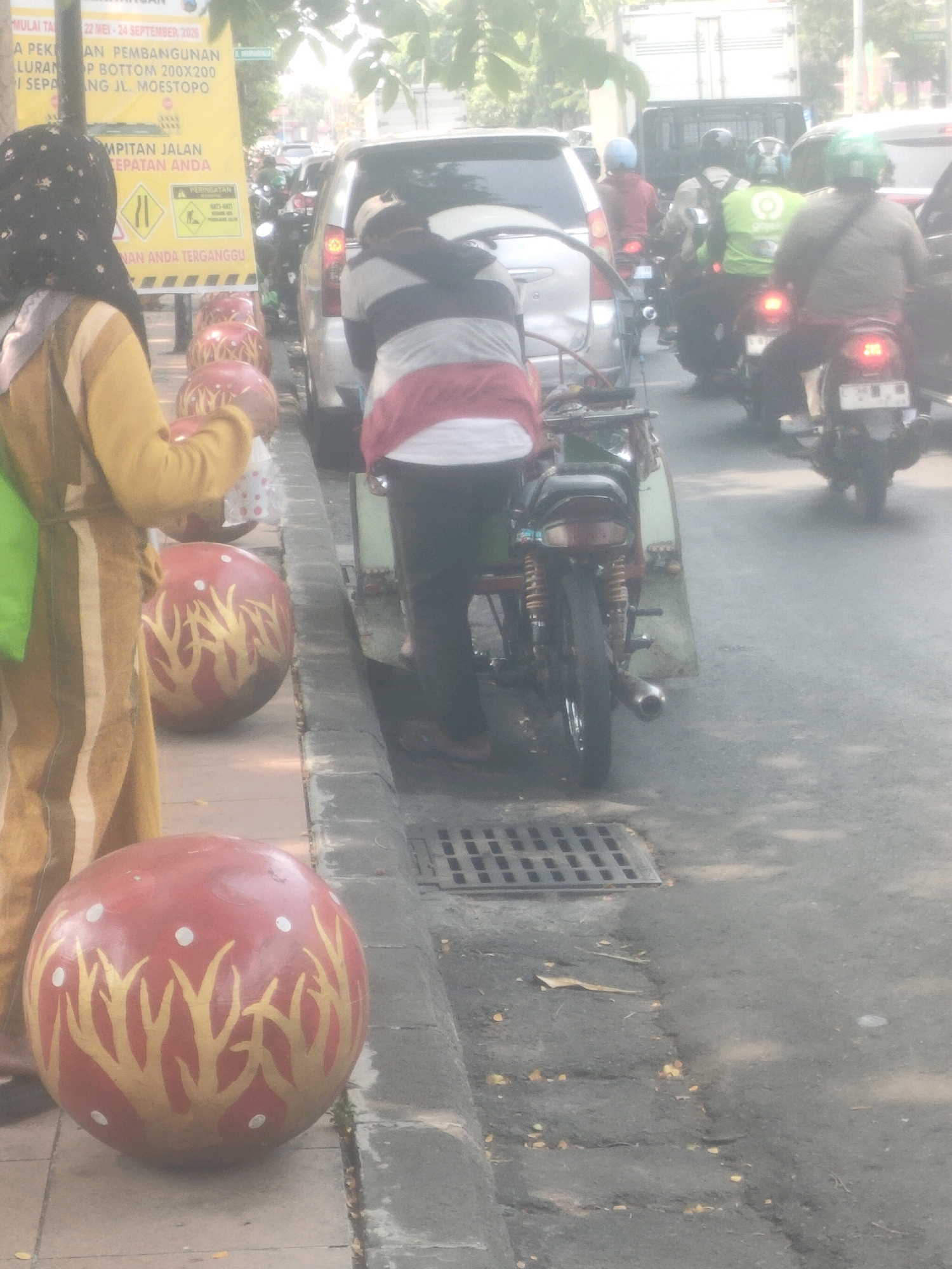 The image depicts a street scene in Surabaya, Indonesia, with a strong emphasis on traffic and pedestrian activity. In the foreground, a woman wearing a yellow tunic and a dark hijab stands to the left, facing right. In front of her are several large, round, red decorative objects with gold tree-like patterns. Behind the woman, a person in a striped shirt and dark pants is seated on a motorcycle, which is part of a three-wheeled vehicle with a wooden side panel.
The background is filled with a variety of vehicles. A silver car is parked on the right, with a truck visible behind it. Several motorcycles are also present, carrying riders wearing helmets of various colors, including green, blue, and red. One of the motorcycle riders is wearing a bright green vest with white lettering. The street itself is asphalt, with a gray concrete sidewalk on the right. A metal grate is visible on the sidewalk.
A yellow banner with text in Indonesian is positioned on the left side of the image, indicating a message related to road construction or maintenance, possibly a warning or announcement. The overall lighting suggests it is daytime, with some haze or glare affecting the clarity of the image. The scene appears to be a busy urban environment, typical of a city like Surabaya.