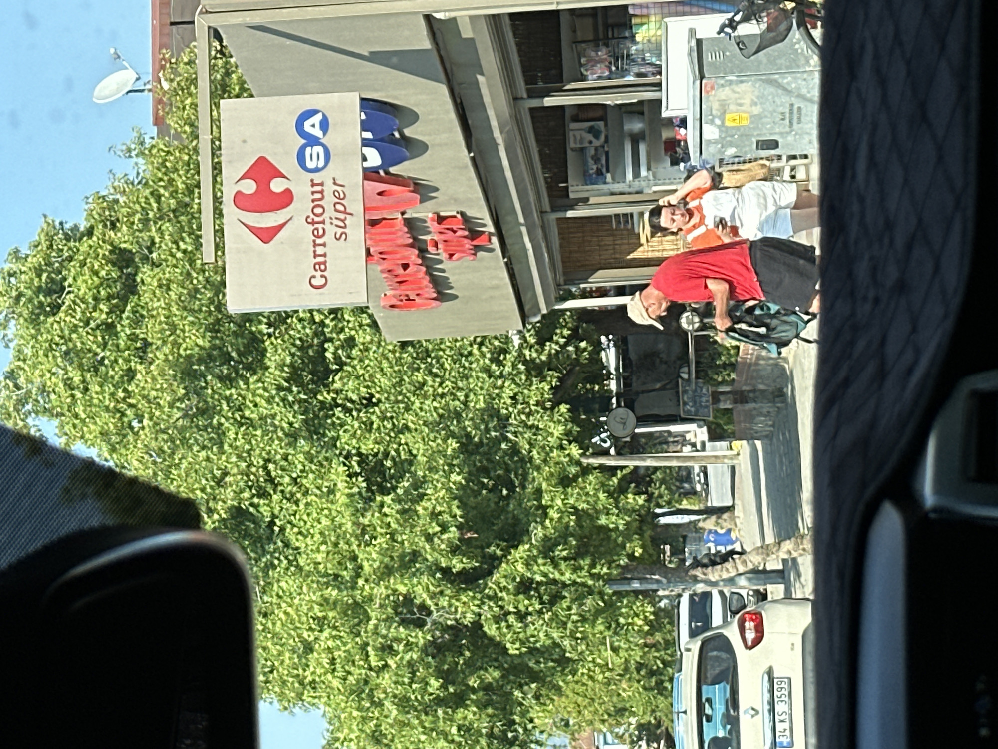 The image, captured from the interior of a vehicle with a dark dashboard visible at the bottom, depicts a sunny street scene in Fethiye, Turkey. On the right, a "Carrefour SA süper" market is prominently featured. The market's main sign displays a red Carrefour logo and "Carrefour SA süper" text; a secondary sign below it also reads "Carrefour Süper" in large red lettering with "SA" in blue. The shopfront consists of glass windows and doors, revealing various household goods and a wooden roll-up blind inside.
Two individuals are present near the market entrance. A man, wearing a red t-shirt, dark shorts, and a light-colored baseball cap, walks towards the left, holding a dark backpack in his left hand. To his right, a woman in an orange t-shirt and white overall shorts stands near the shop, looking down at a mobile phone in her hand, with her left arm raised towards her head. Adjacent to the shop entrance, a gray electrical box with yellow warning labels is visible.
The street is lined with abundant, lush green trees, creating a dense canopy under a clear blue sky. On the left side of the image, a white Renault Clio car, displaying the Turkish license plate "34 KS 3599," is parked facing away from the viewer. Another white vehicle is partially visible behind it. In the upper right background, a white satellite dish is mounted on a building rooftop. Further down the street on the right, additional trees and a pole with a partially obscured round sign are visible.